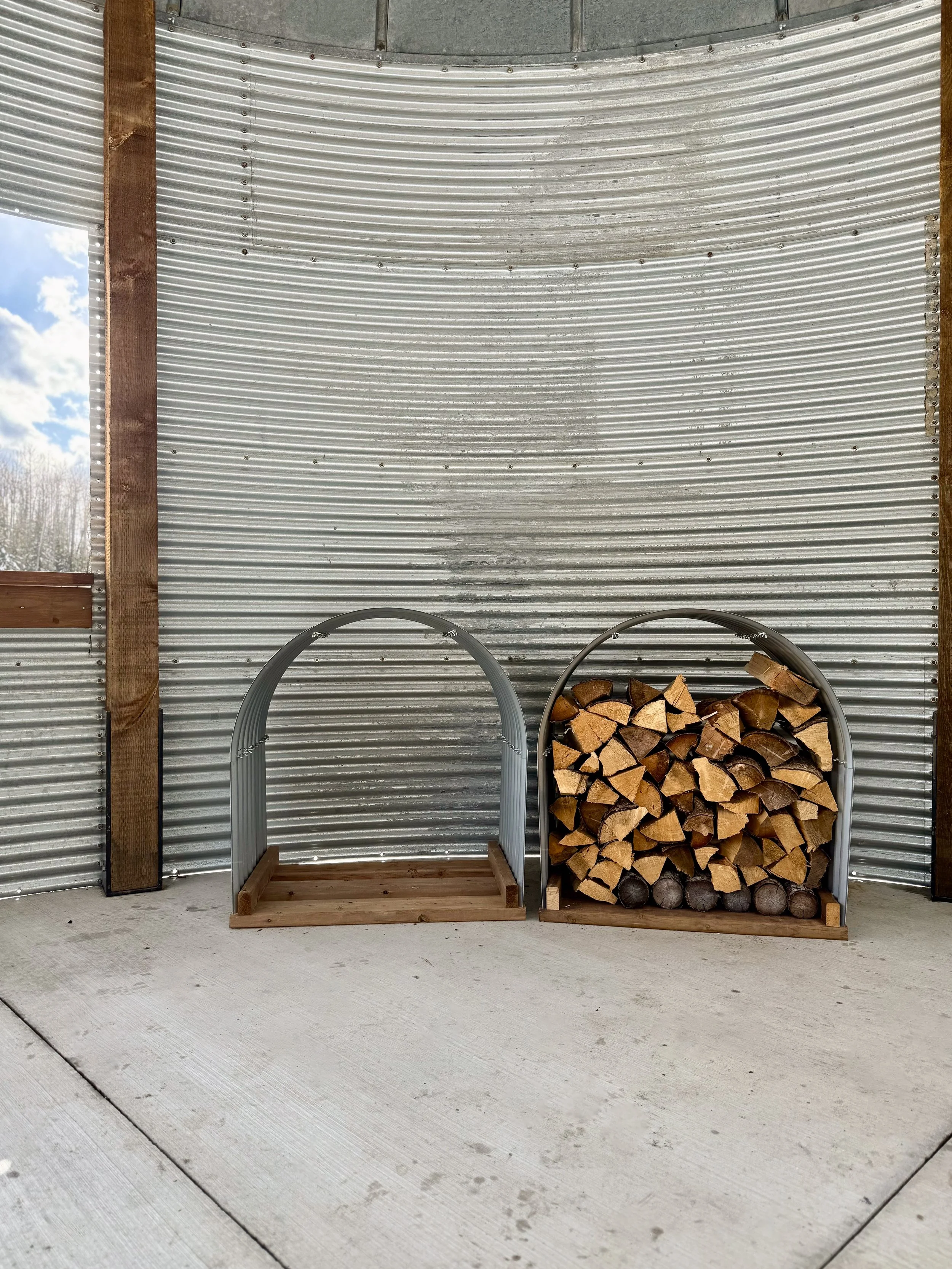 Two metal firewood racks, one empty and one filled with stacked firewood, against a corrugated metal wall.