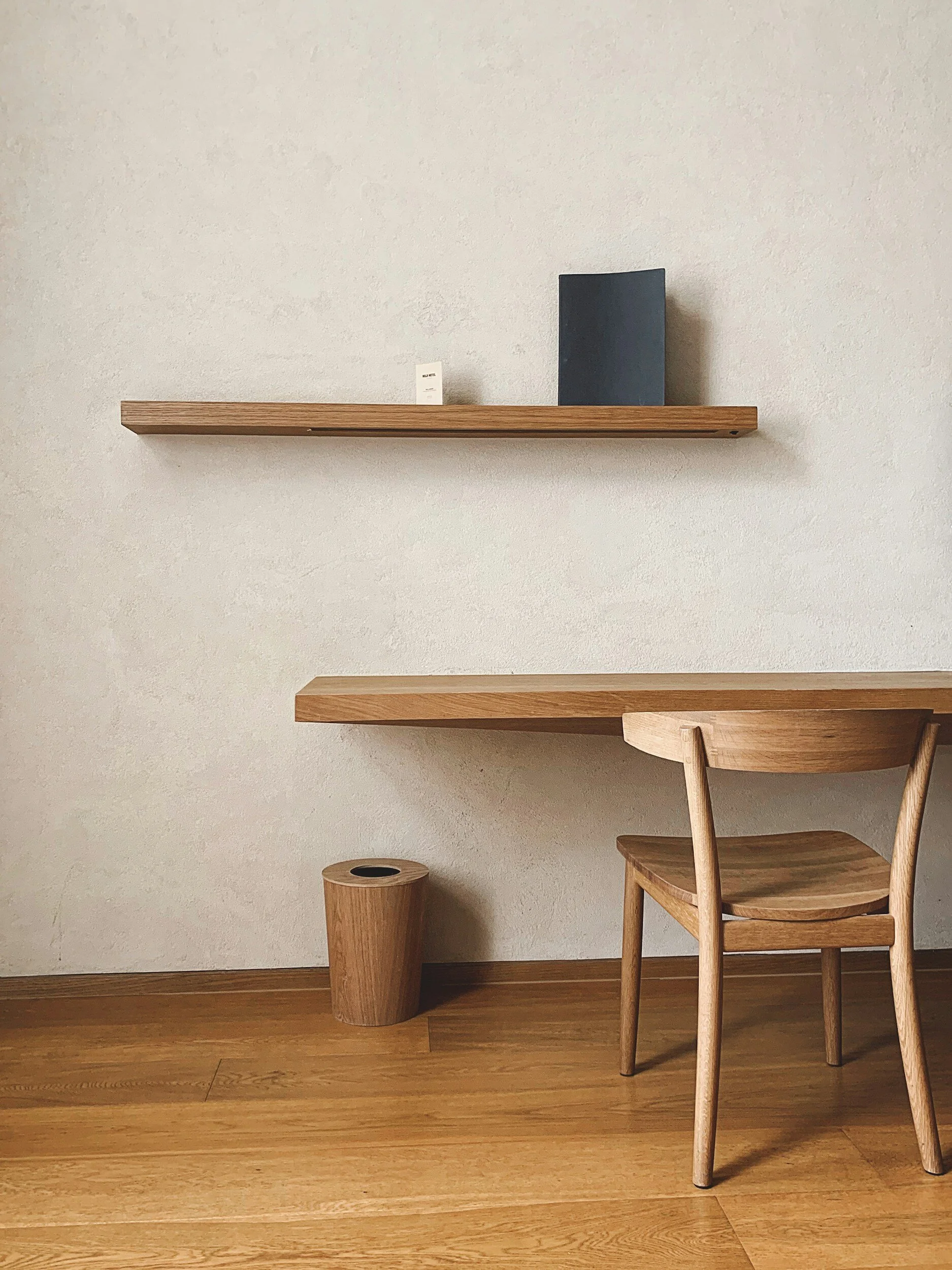 Minimalist wooden desk with a matching wooden chair, a wood wastebasket, a floating wooden shelf with a small book and a dark-colored notebook, against a neutral-colored wall and wooden floor.