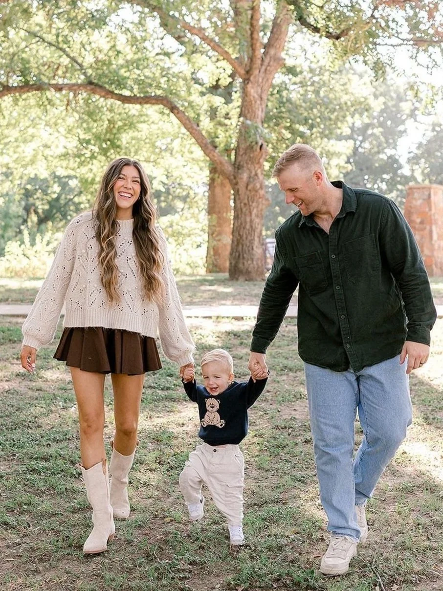 A happy family of three walking outdoors in a park, holding hands and smiling, with trees in the background.