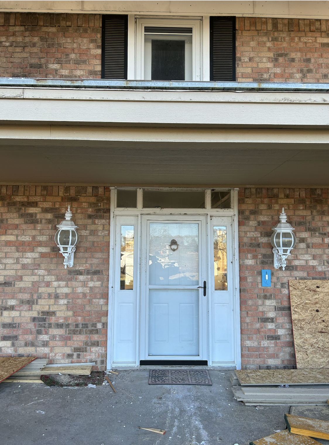 Front entrance of a house under renovation with a white door, brick walls, and two white lantern-style light fixtures on either side. Plywood boards are leaning against the wall, and construction debris is on the ground.