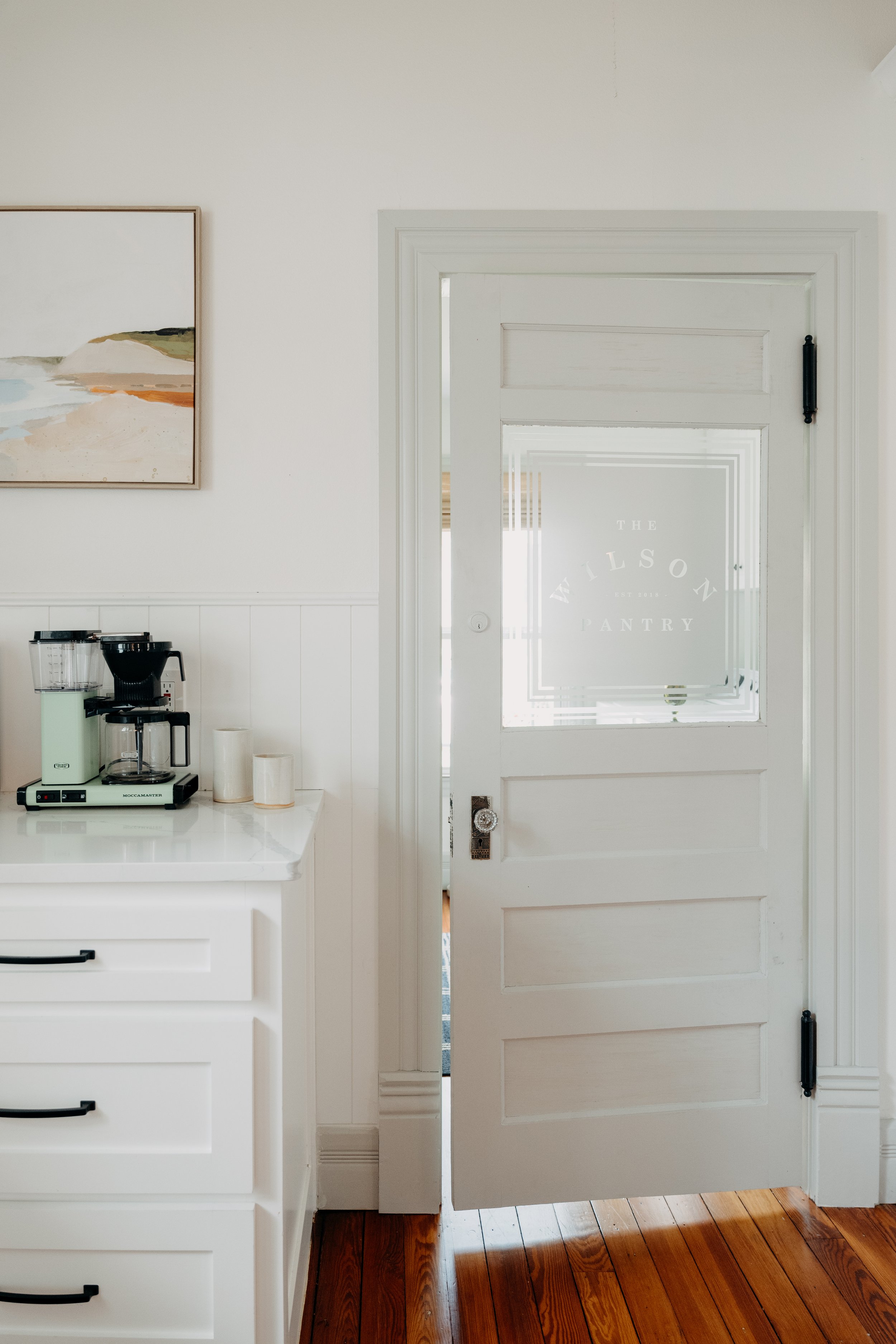 White door with glass window labeled 'The Wilson Pantry' in a bright kitchen with wooden floors and white cabinetry.