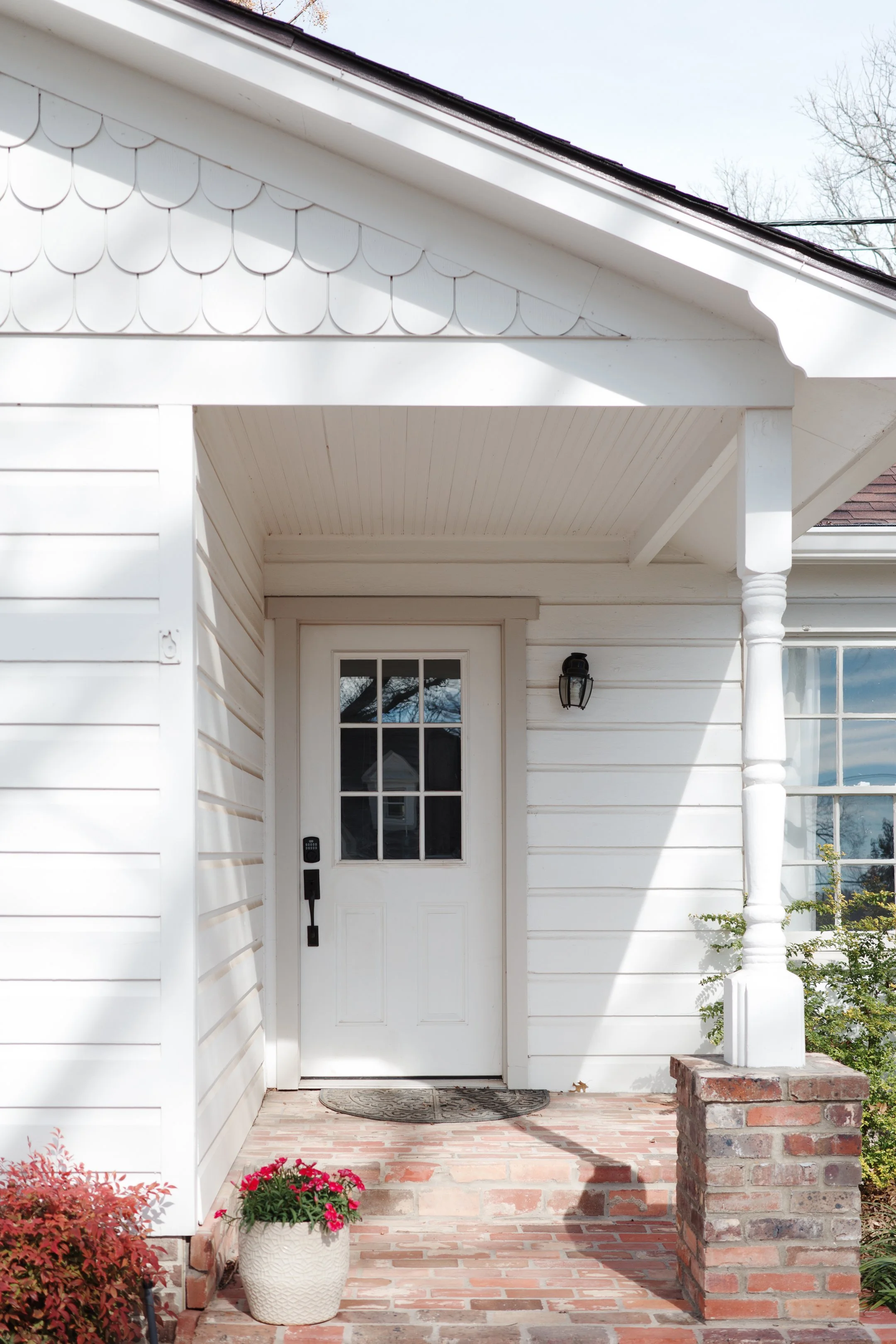 White house exterior with a front door, brick pathway, potted flower, and porch light.