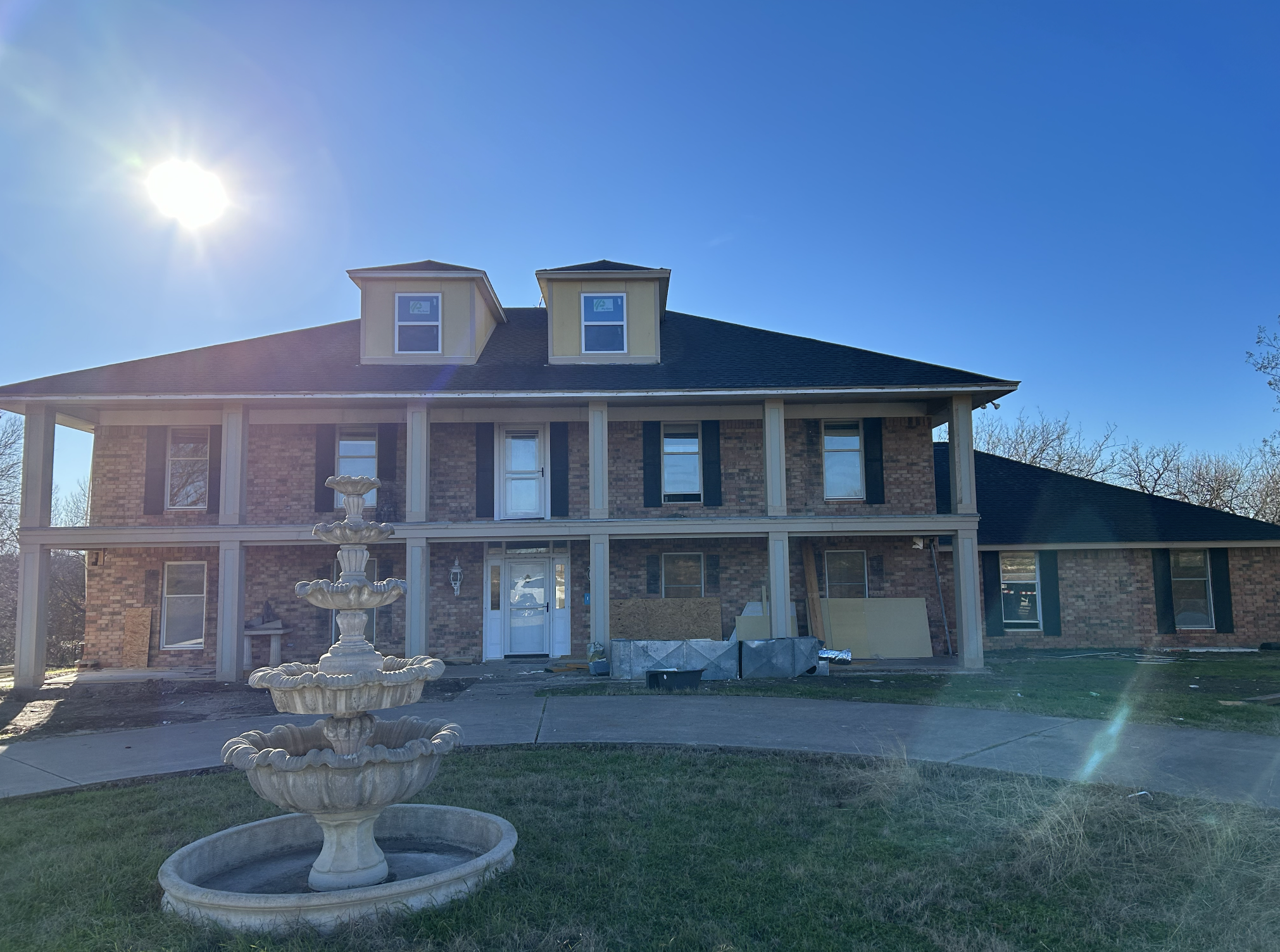 A large brick house under construction with scaffolding at the front and a decorative fountain in the yard. The house has two stories, several windows, and dormer windows on the roof. The sky is clear and the sun is shining brightly.
