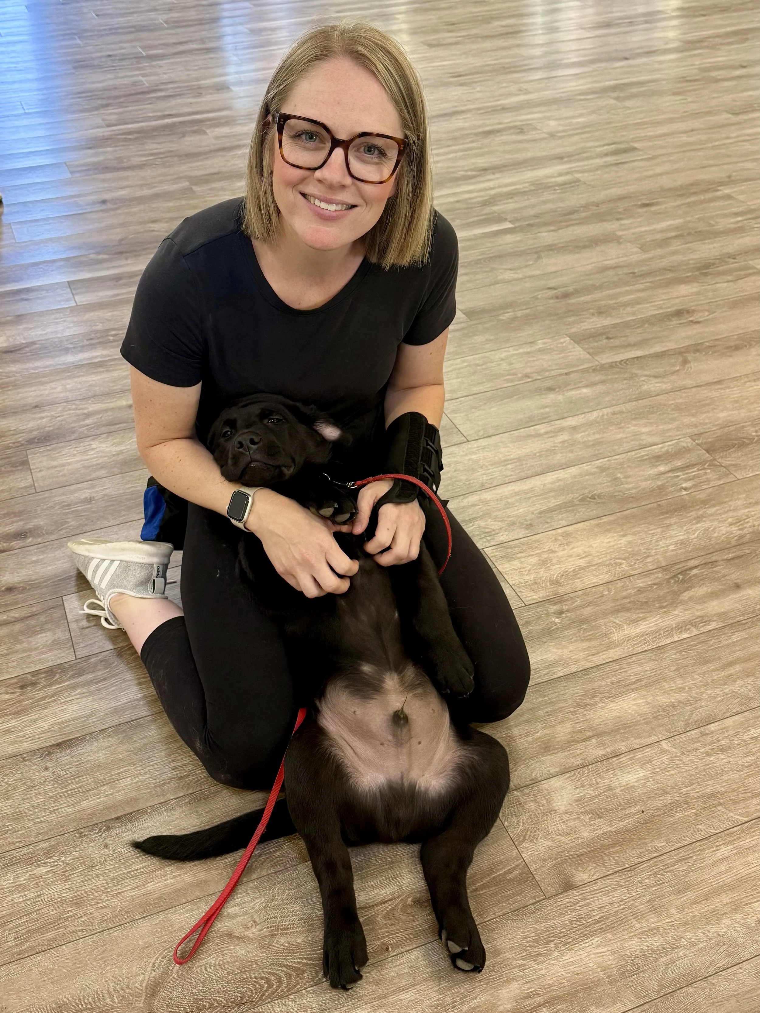 A woman with short blonde hair, glasses, and a black shirt sitting on a wooden floor, holding a black puppy, smiling at the camera.