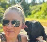 A woman wearing sunglasses smiling while a black dog licks her face outdoors on a sunny day.