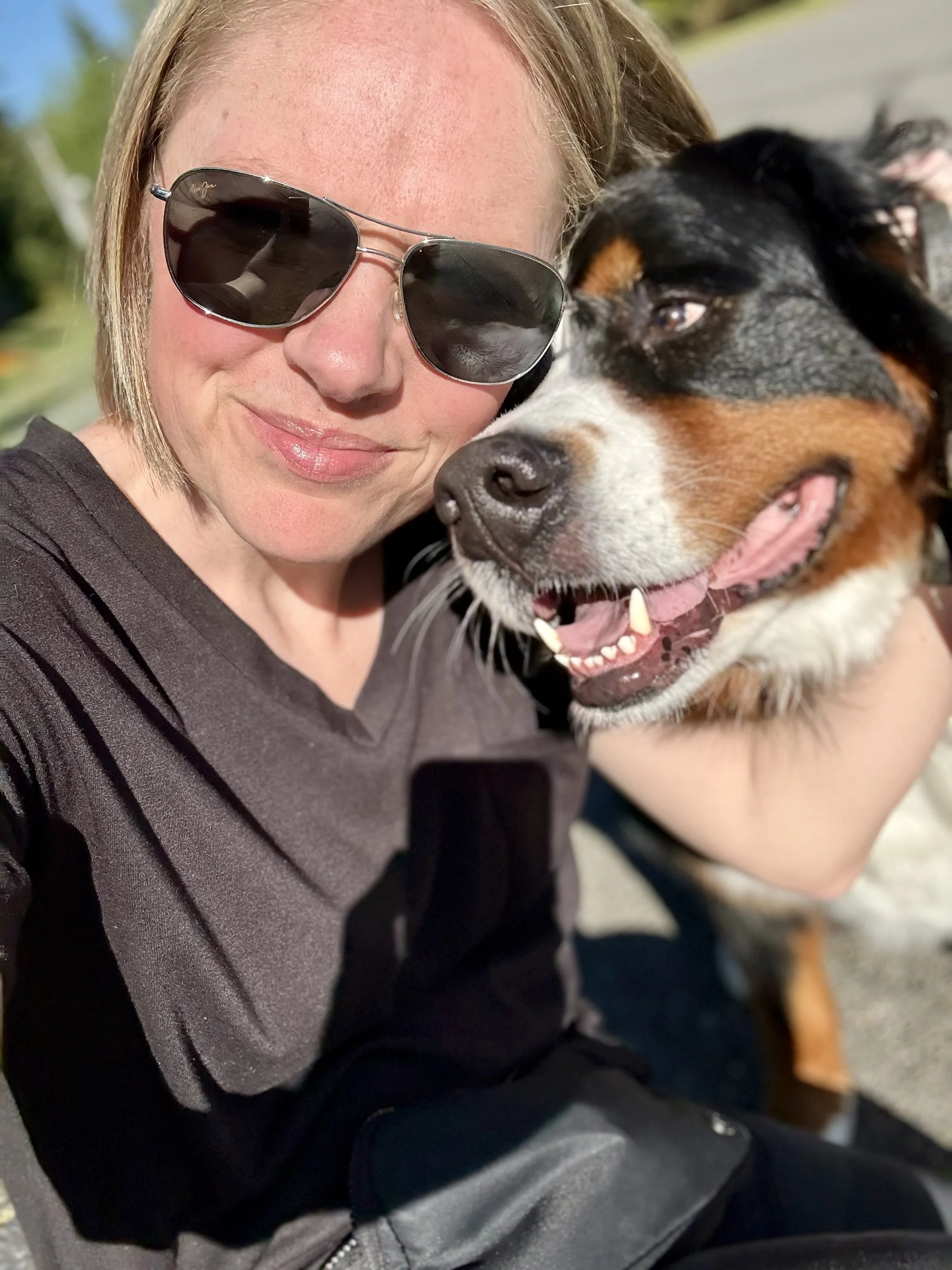 A woman wearing sunglasses and a dark shirt, smiling, holding a large dog with black, brown, and white fur, and the dog's mouth open with a pink tongue visible.