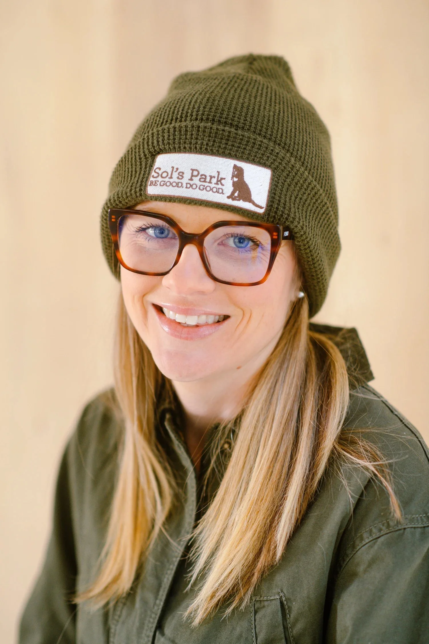 A woman with long blonde hair wearing glasses and an olive green beanie hat with a patch that says Sol's Park, BE GOOD. DO GOOD.