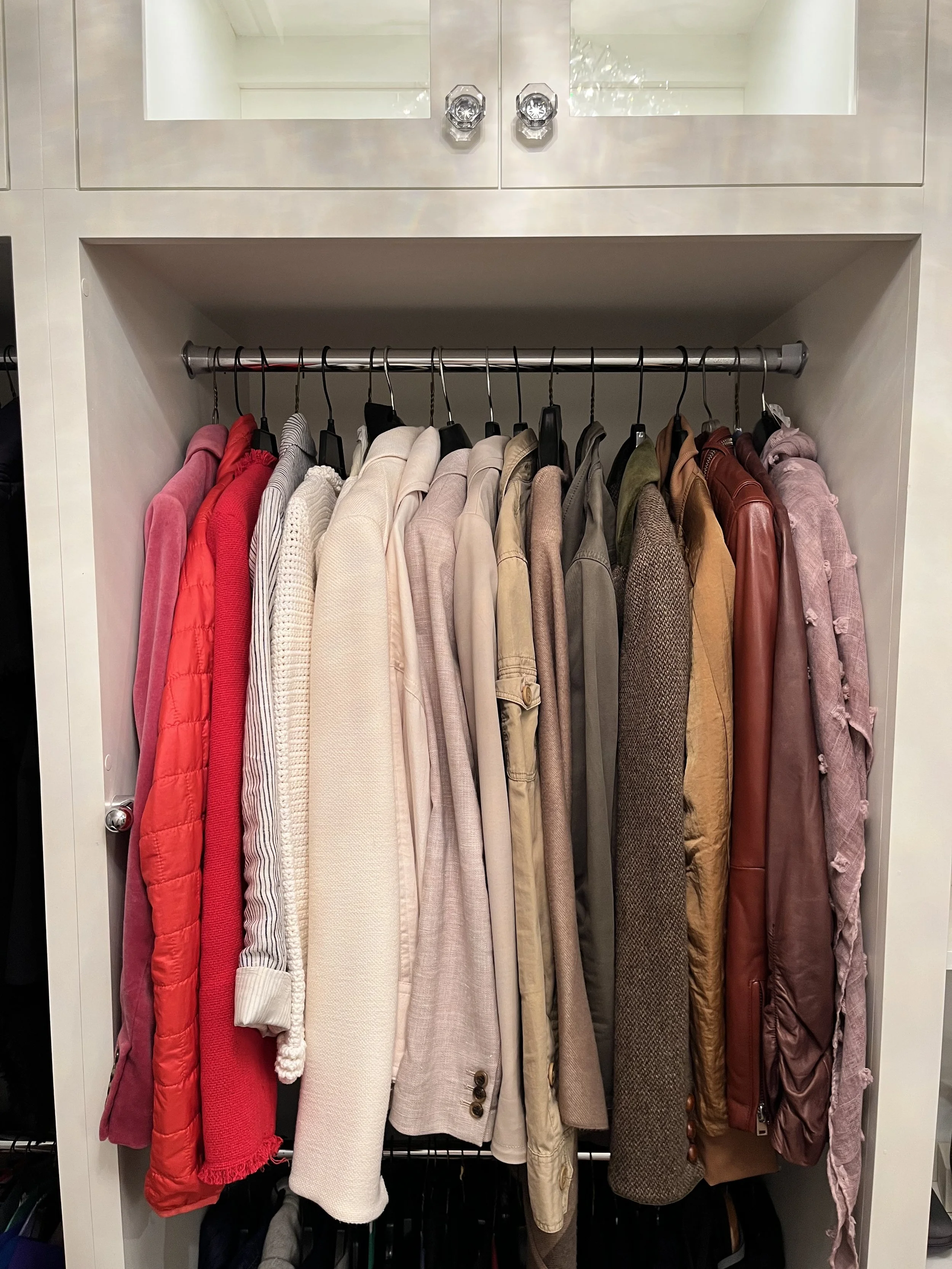 An organized closet with color coded coats hanging on a metal rod, surrounded by a built-in shelf and cabinet. This Atlanta closet organization is by Happy Tree Organizing, based in Decatur, GA. 