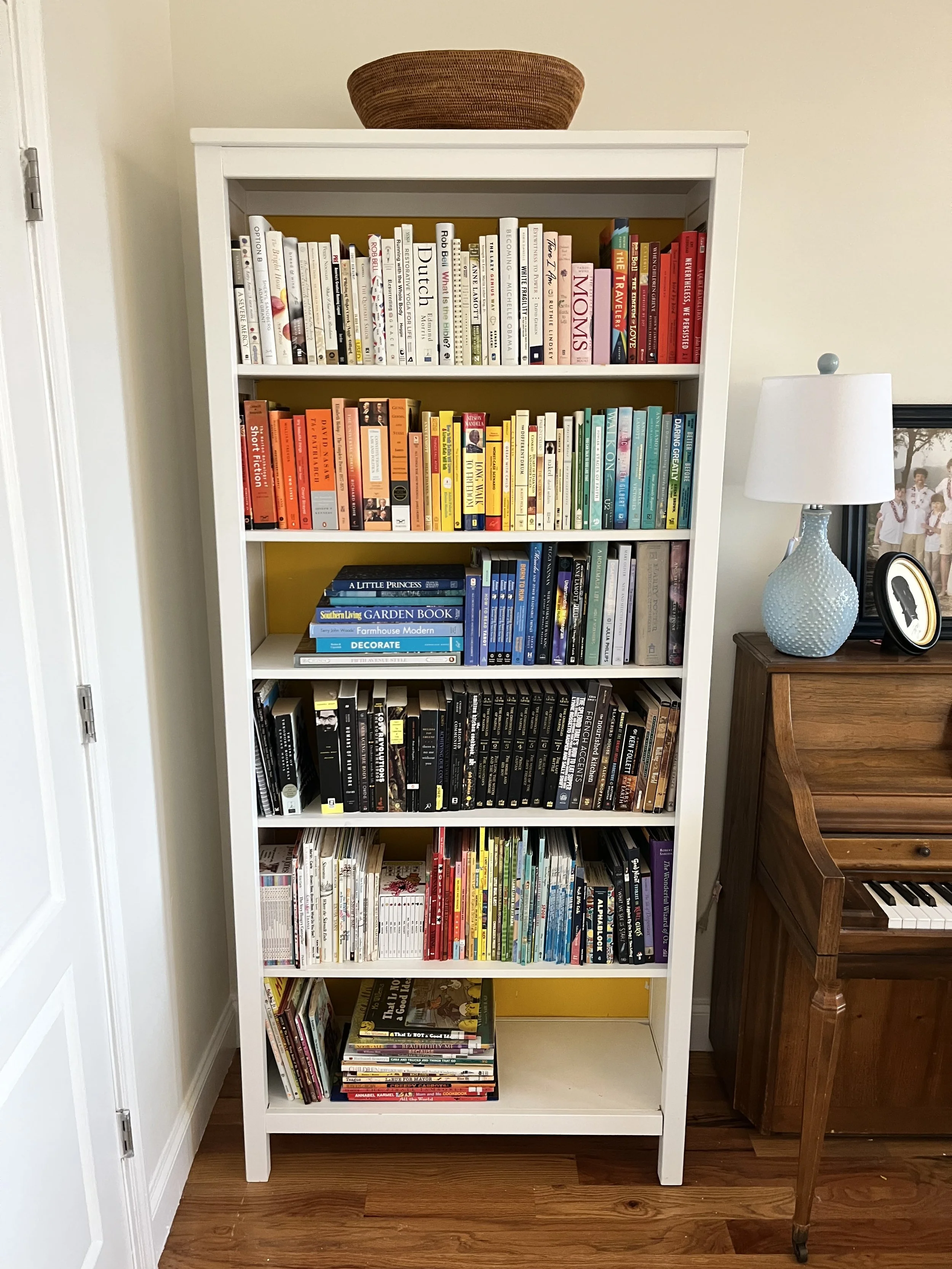 Styled and organized bookshelf in Decatur, GA, filled with colorful books, next to a wooden piano with a lamp and framed photograph on top, against a beige wall. 