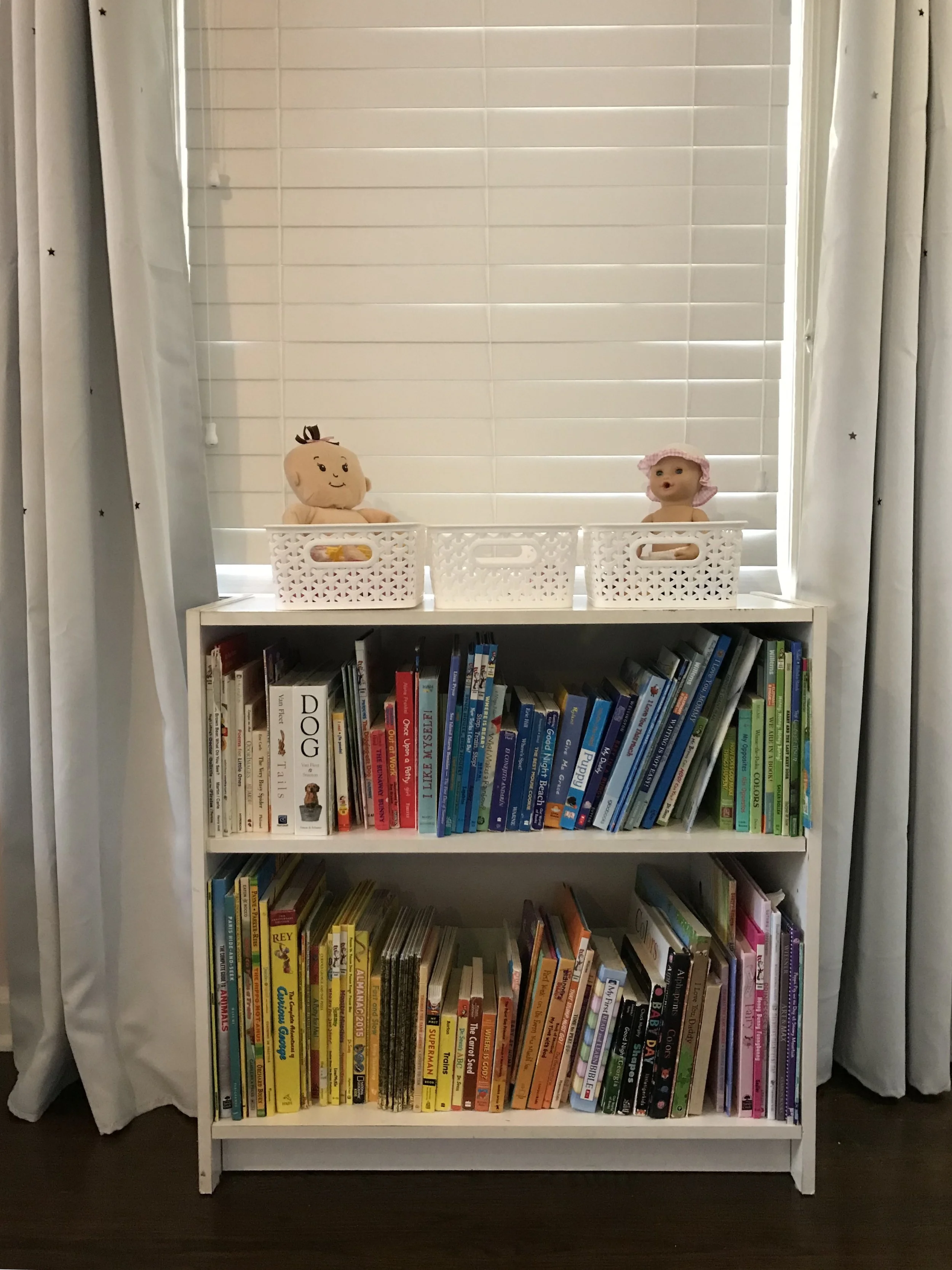 Kids organization tips are shared here, by Happy Tree Organizing. A white bookshelf filled with organized children's books, topped by adhd friendly organization - open top white baskets, in front of white blinds  curtains on either side.