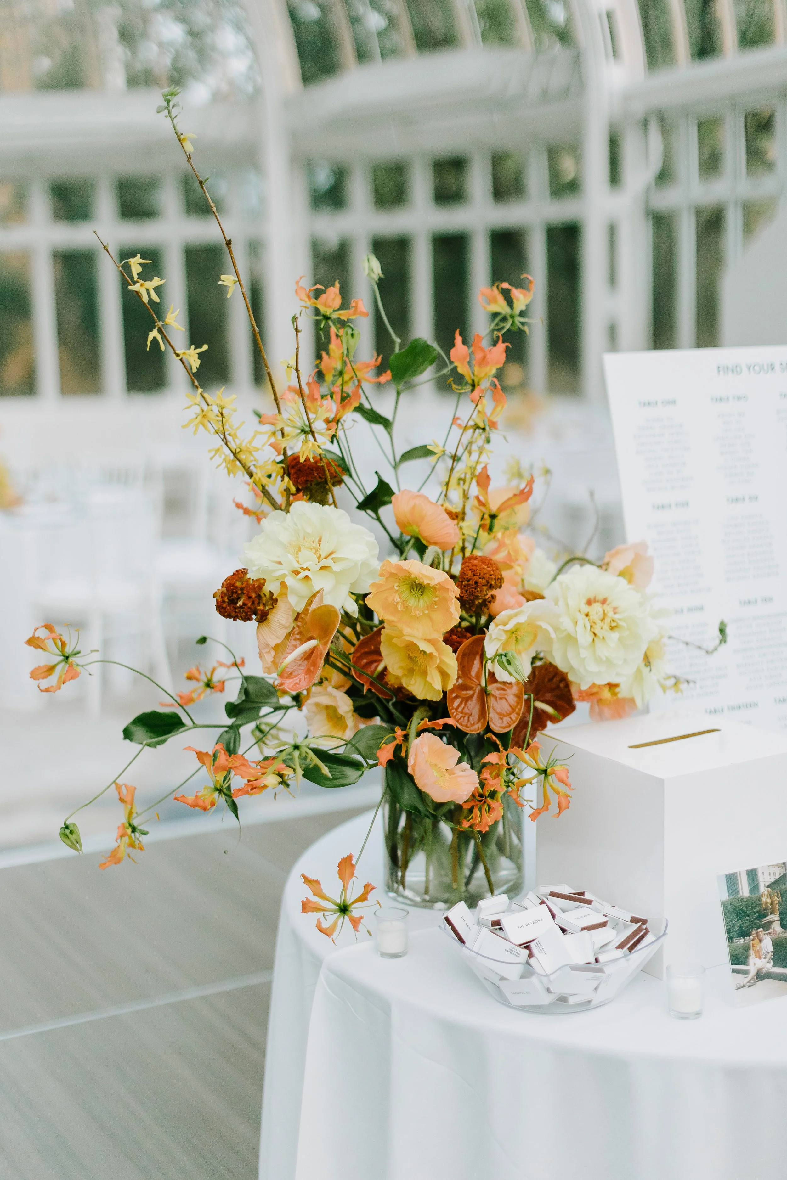 A floral arrangement in a glass vase with peach, white, and green flowers on a white table. There are small votive candles, a bowl of small boxes, and a large white card or menu on the table. The background shows a window with a lattice design.
