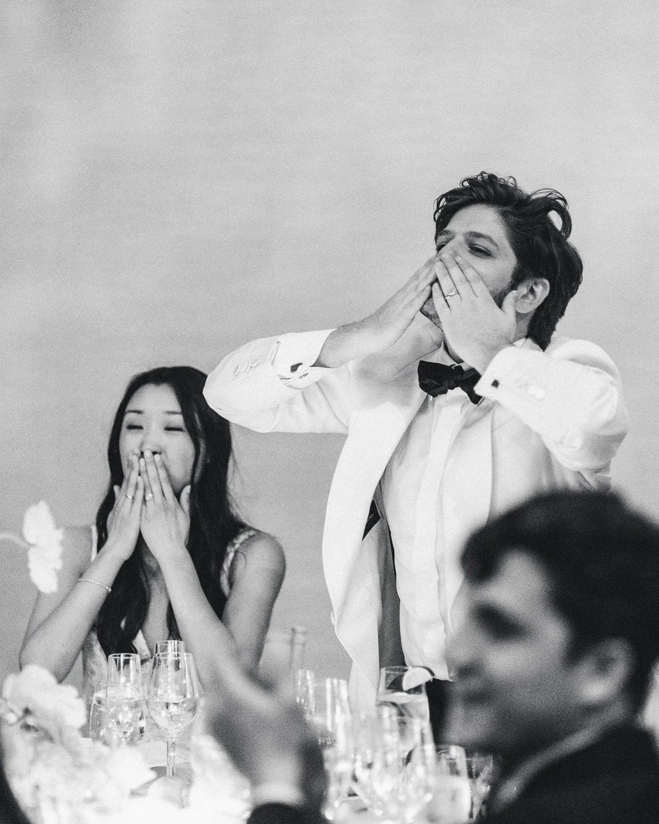 Black and white photo of a man in a tuxedo and a woman dressed nicely at a formal event, both covering their mouths with their hands, appearing to be surprised or emotional.