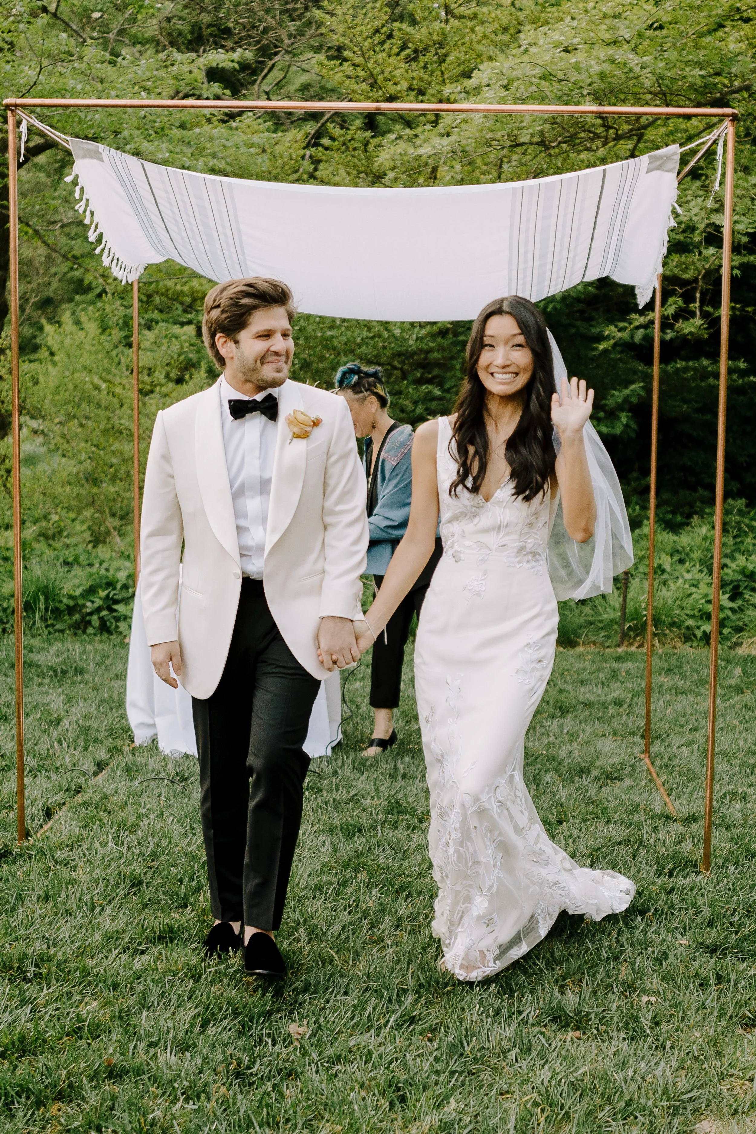 A newlywed couple walking hand-in-hand outdoors on a grassy lawn, the bride waving and smiling, with a white canopy and green trees in the background.