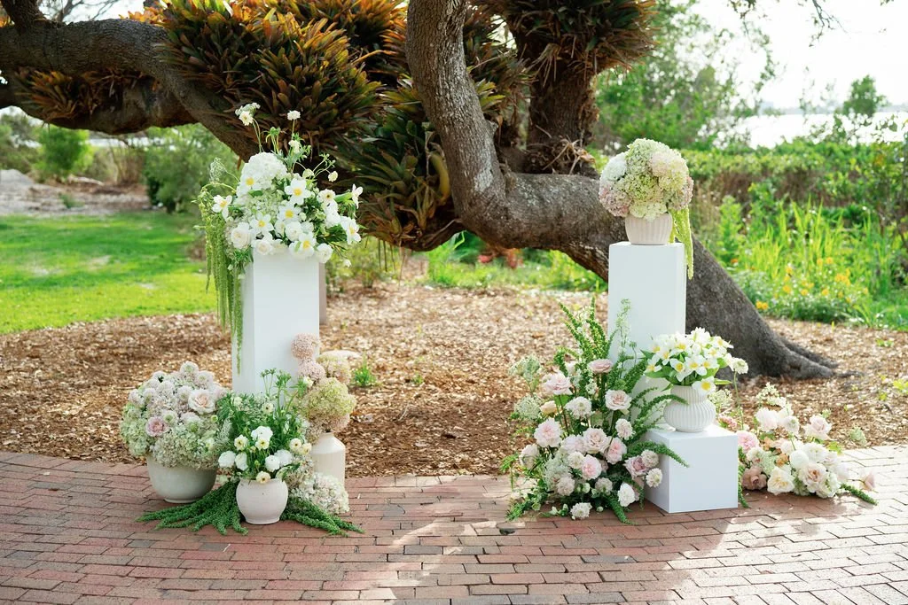 Outdoor floral display with white and pink flowers, vases, and white pedestals under a large tree.