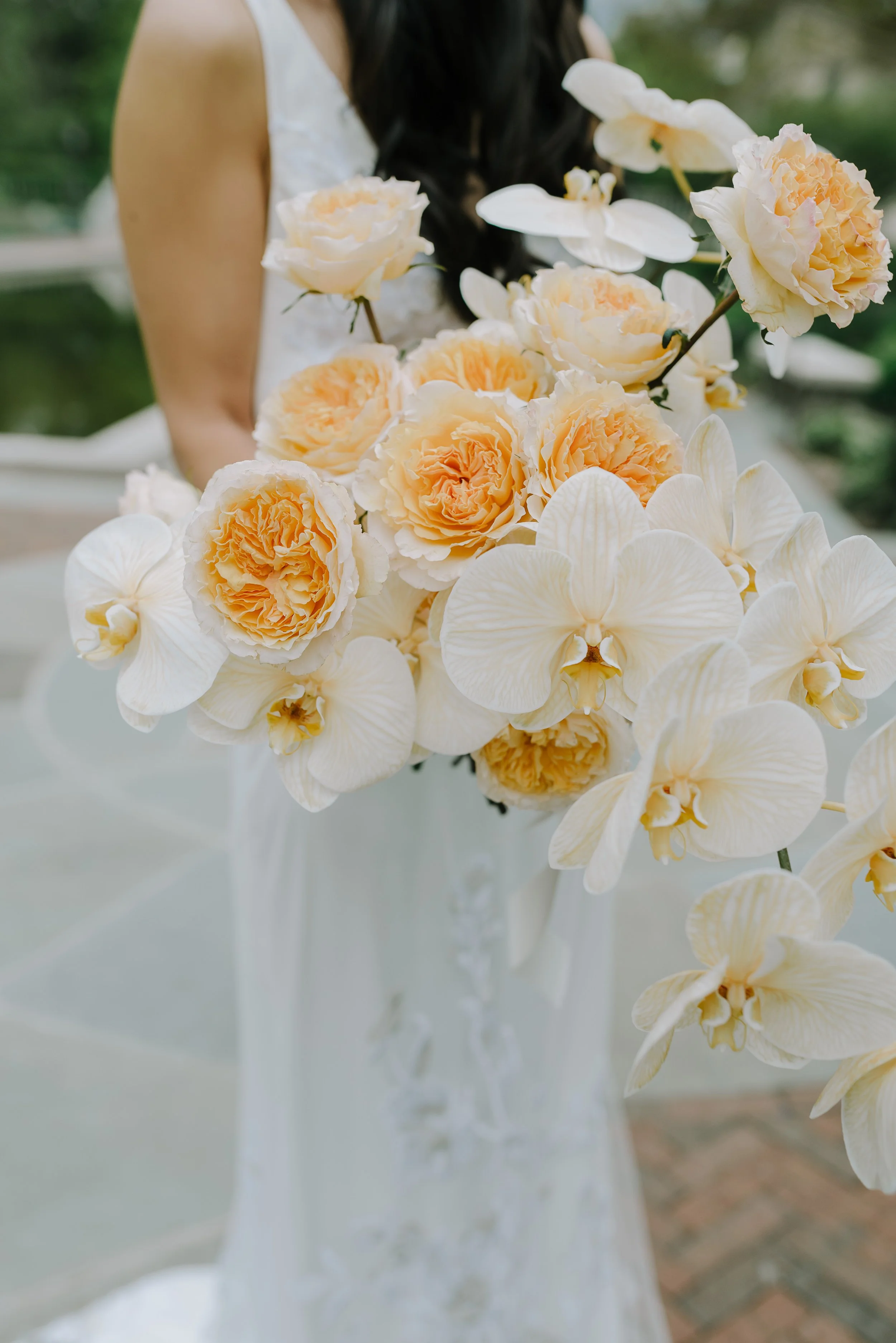 A person holding a large bouquet of cream-colored orchids and peach peonies, with a lace wedding dress visible. The background is blurred greenery.