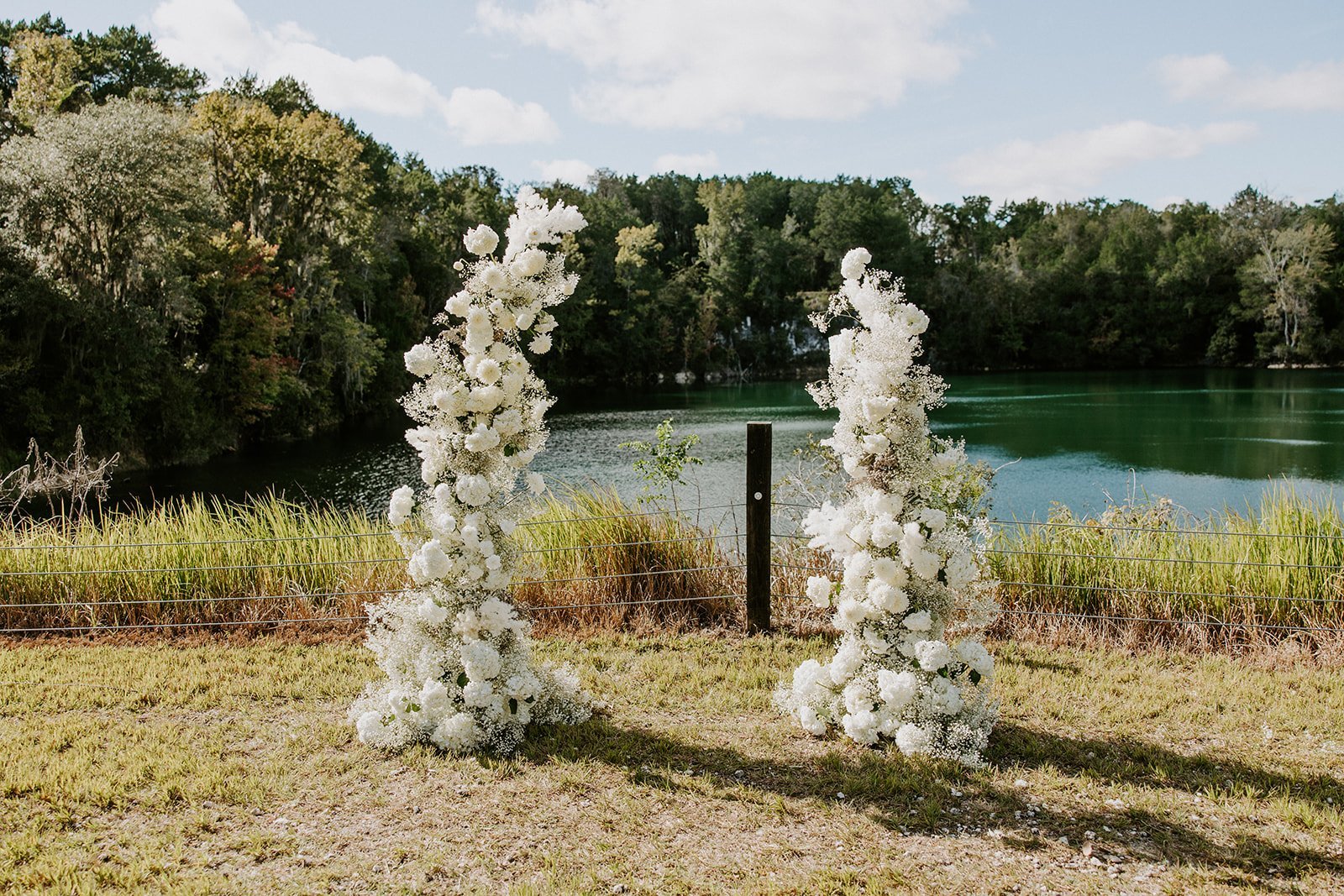 Two tall floral arrangements made of white flowers are placed outdoors near a lake, with trees and blue sky in the background.