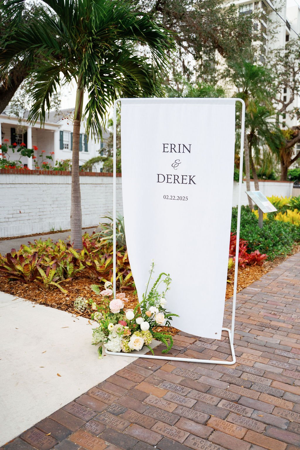 Wedding sign with the names Erin and Derek, the date 02.22.2025, decorated with a bouquet of white and pink flowers, in an outdoor garden setting with trees and plants.