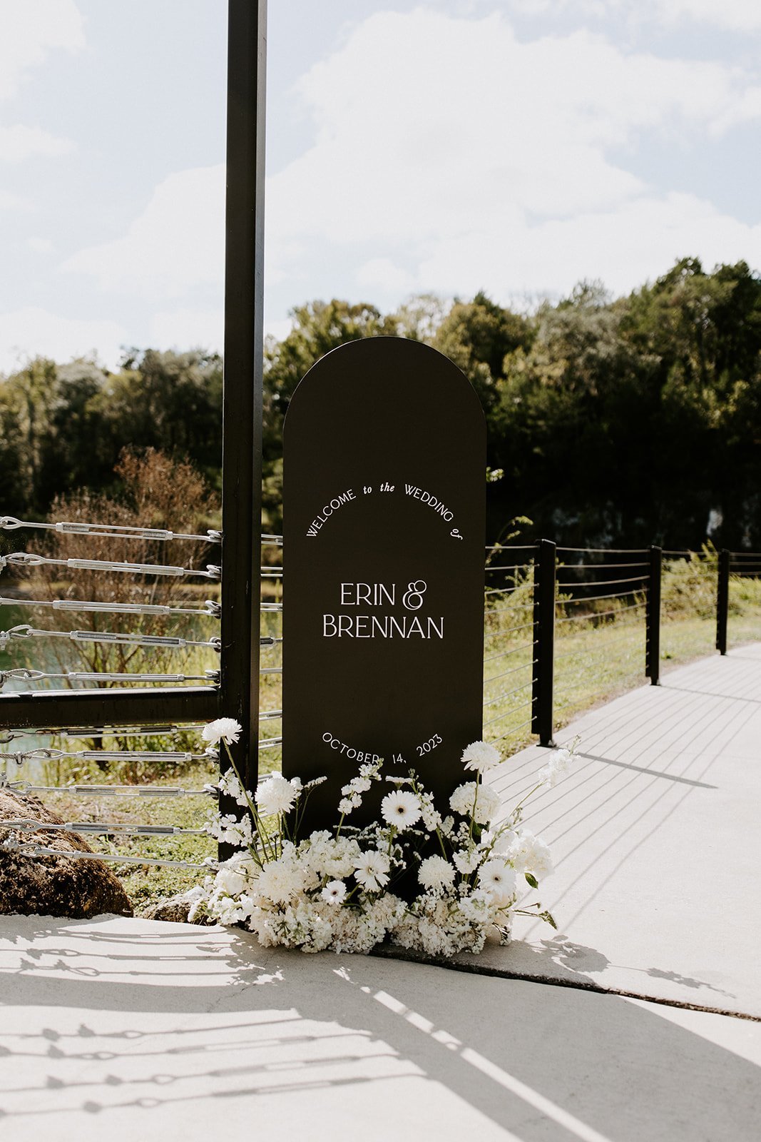 Black wedding sign with white text, decorated with white flowers, indicating the wedding of Erin and Brennan on October 14, 2023, outdoors with a metal railing and trees in the background.