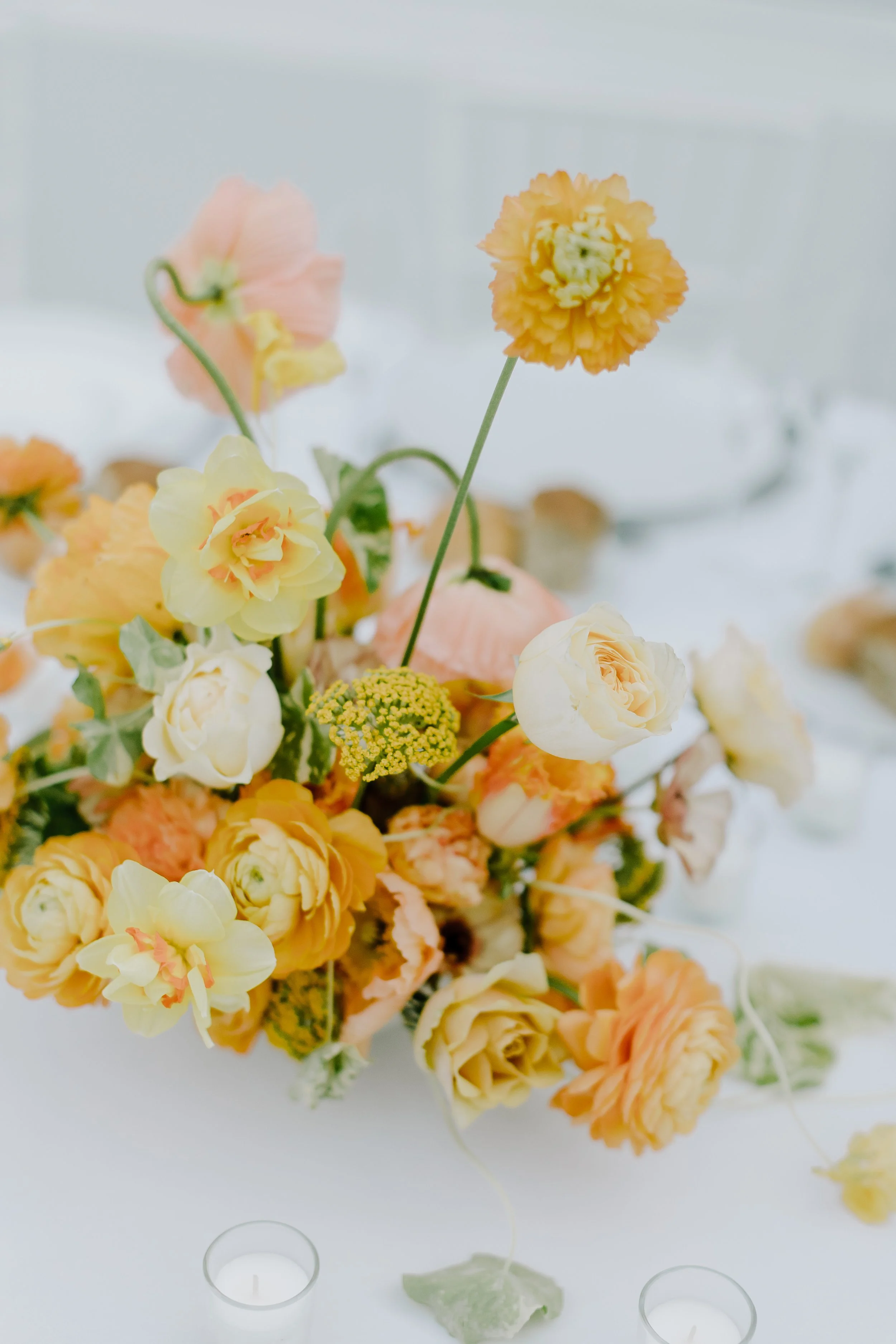 A bouquet of yellow, peach, and cream roses, ranunculus, and carnations arranged on a white surface, with small tealight candles surrounding it.