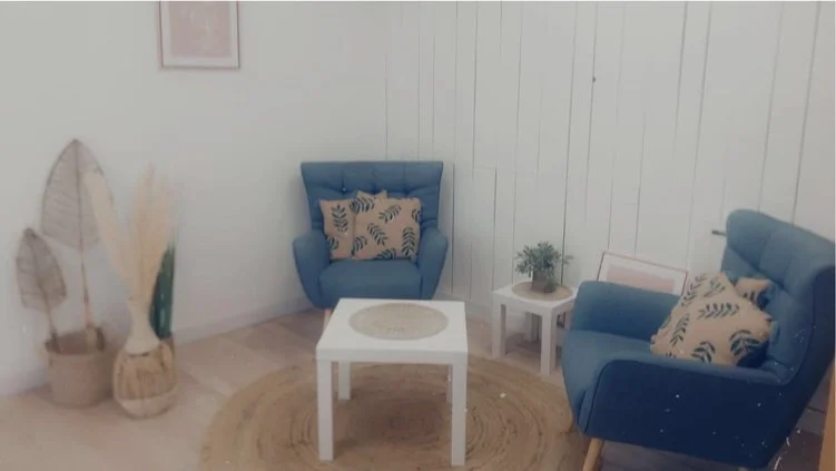 Salon avec deux fauteuils bleus, une table blanche, des coussins à motifs, une plante, et des décorations naturelles dans un style minimaliste.