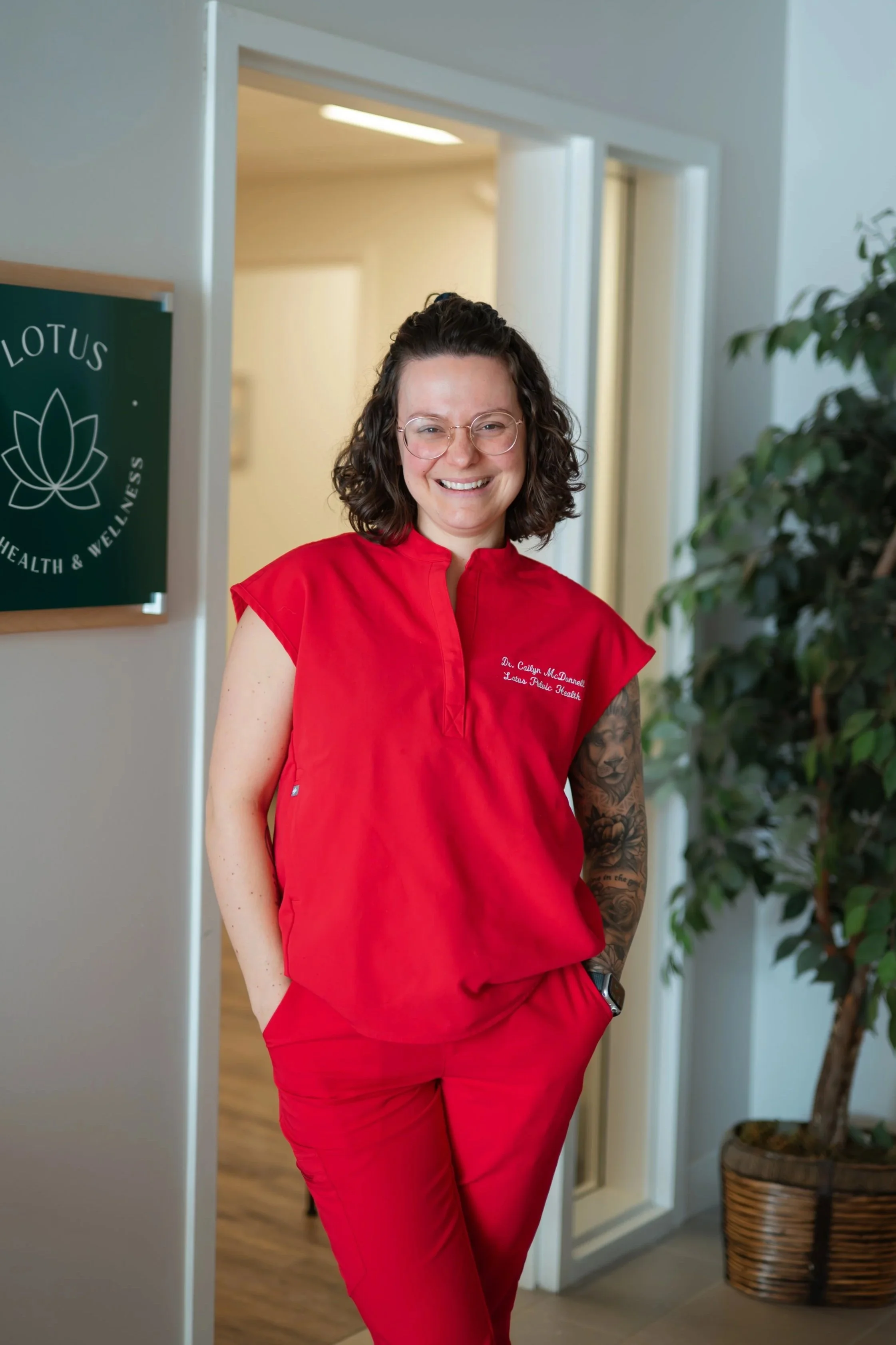 A woman in red medical scrubs standing in a wellness clinic, smiling with hands in pockets, in front of a doorway with a plant nearby.