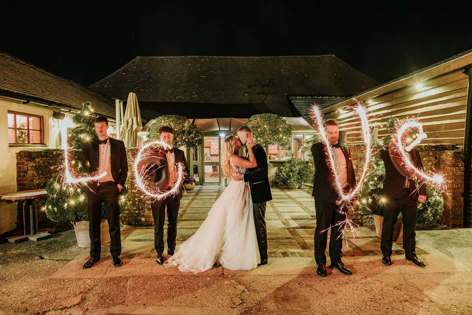 Wedding couple kissing in the center surrounded by four men in tuxedos holding sparklers that spell out 'LOVE' at night.