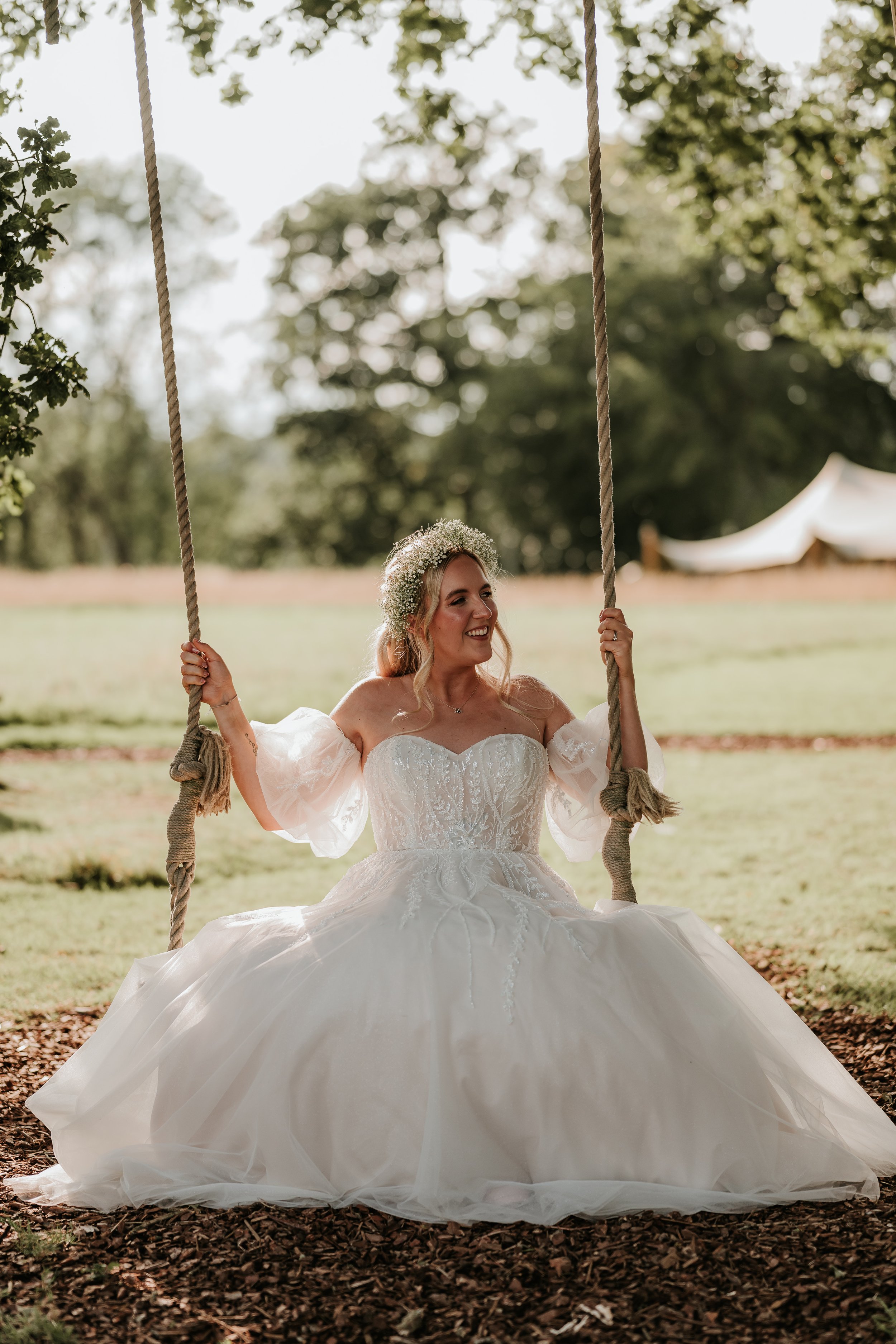 Bride in a wedding dress sitting on a swing outdoors, smiling with a flower crown in her hair.