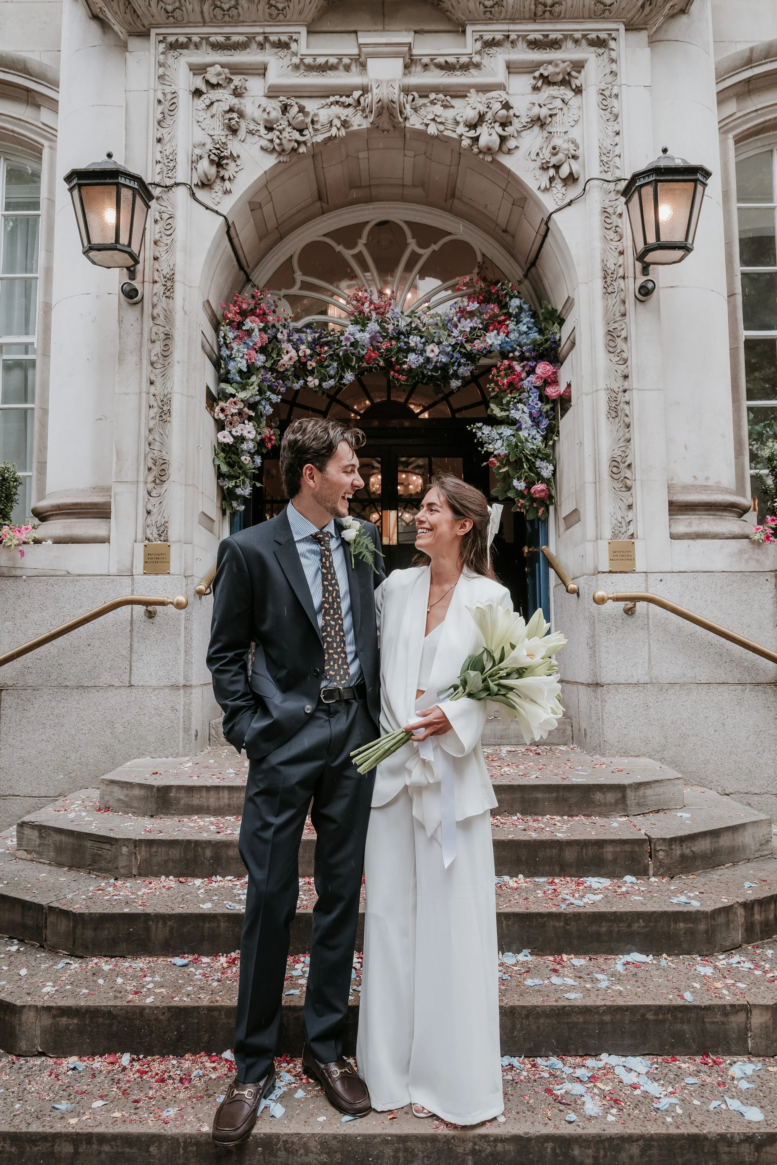 A couple in wedding attire standing on the steps of a building with floral decorations, holding a bouquet of white lilies, smiling at each other.