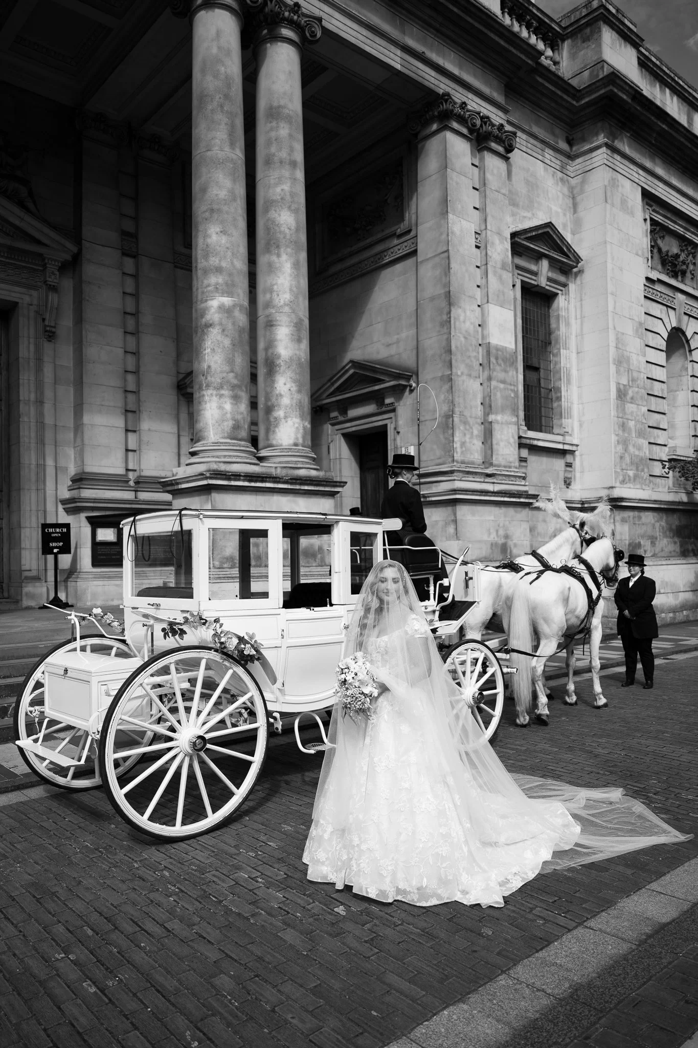 Black-and-white photo of a bride in a wedding gown and veil standing next to a white horse-drawn carriage in front of a large stone building with columns.