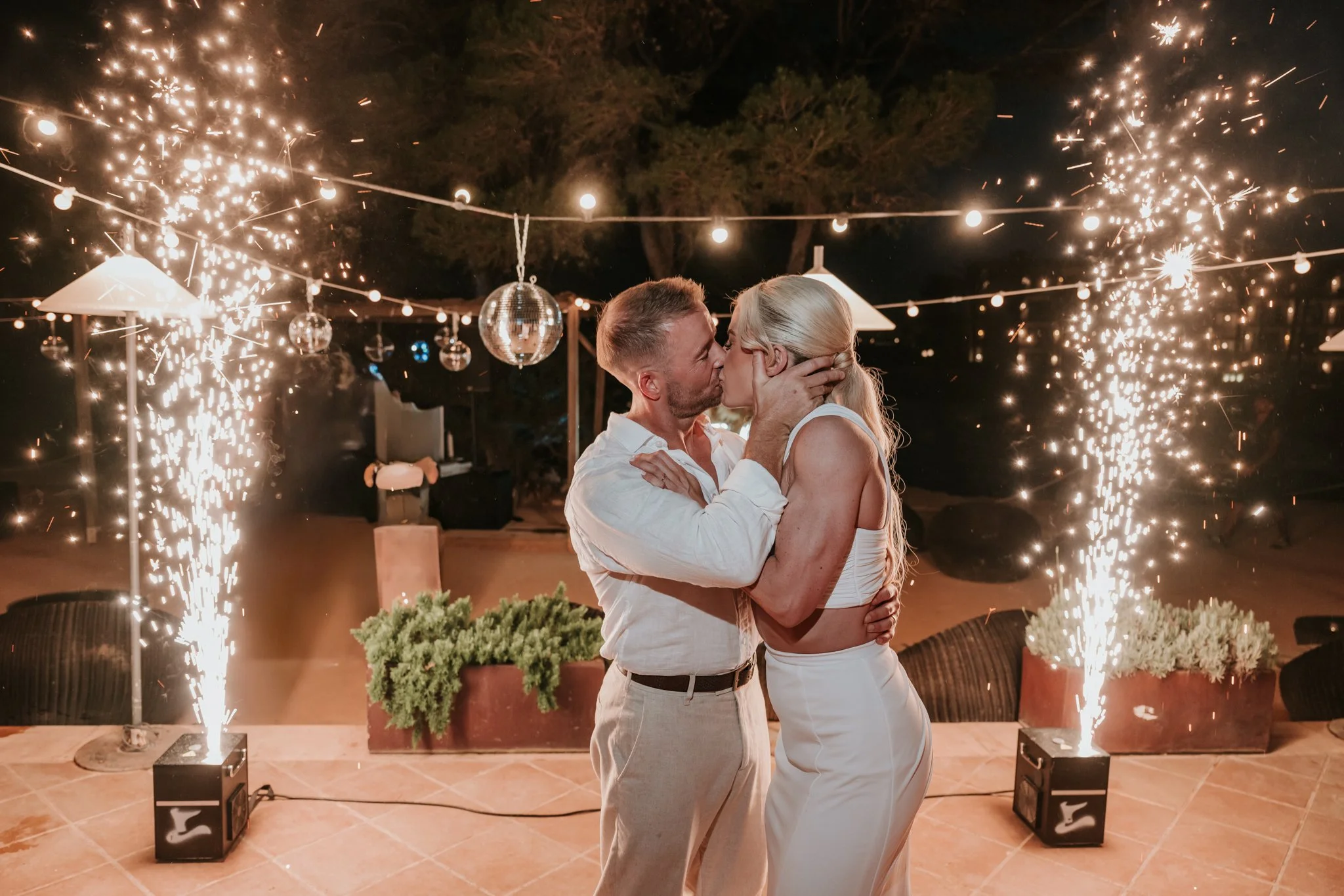 A couple sharing a kiss at a celebration with sparklers and disco balls hanging above them at night.