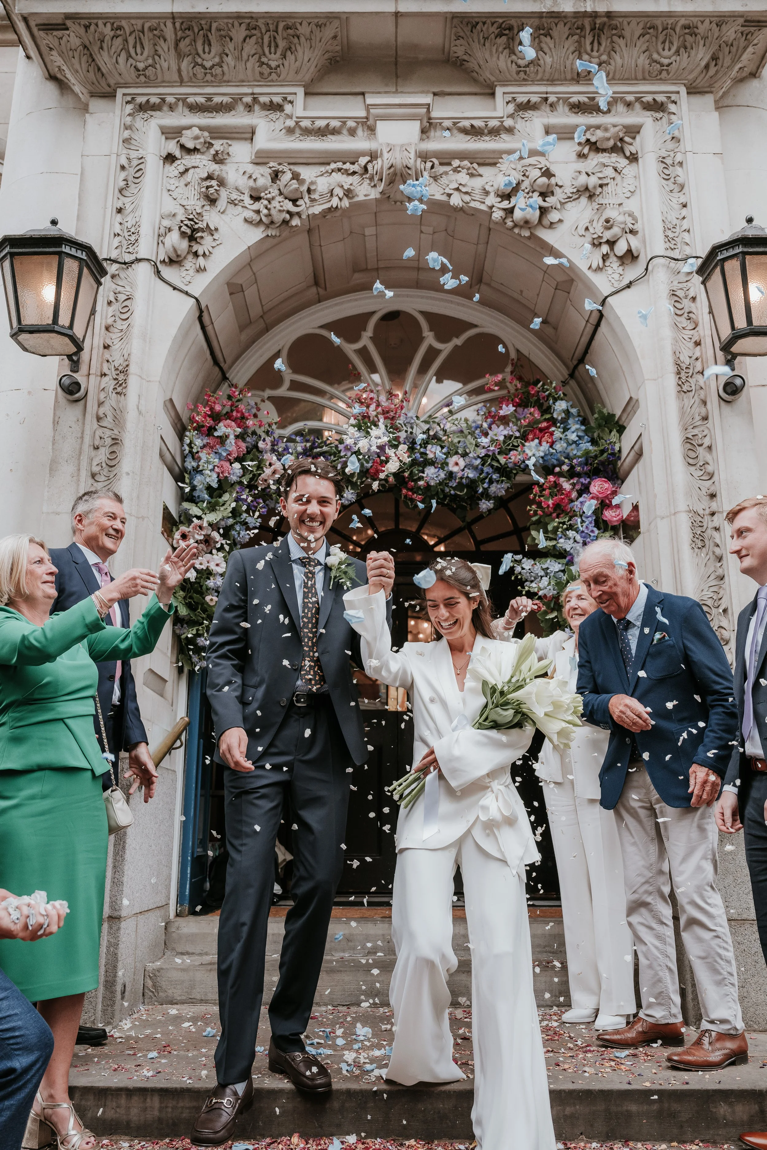 A newly married couple celebrating with family as they exit a building decorated with flowers, confetti, and blue paper butterflies.
