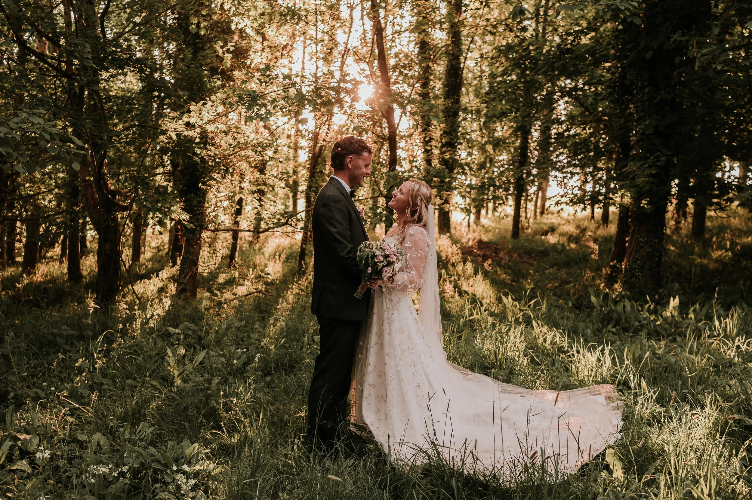 A bride and groom stand face to face in a forest during sunset, holding hands and smiling at each other. The bride wears a long white wedding dress with lace details and a veil, and holds a bouquet of flowers. The groom wears a dark suit. Sunlight fi