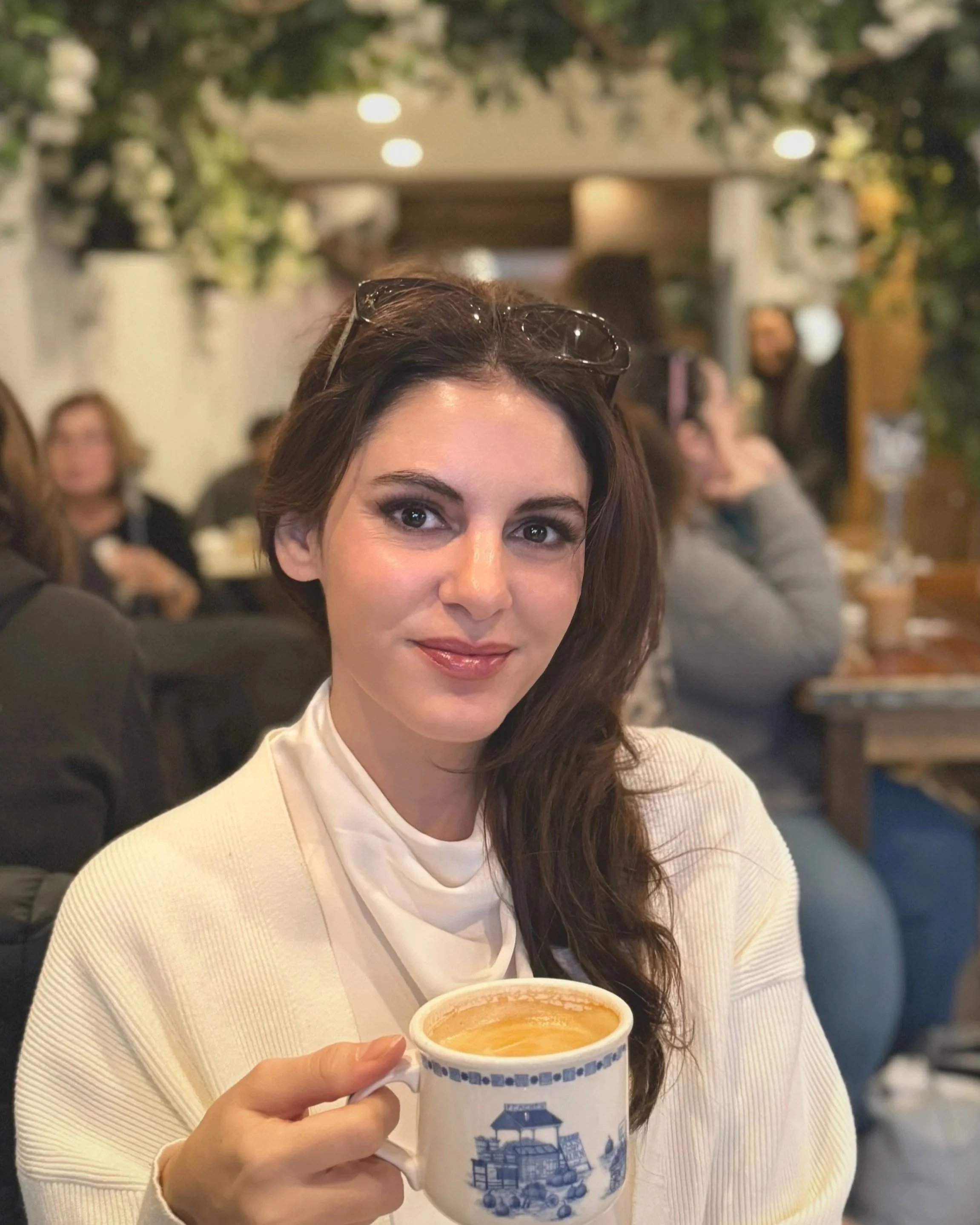 A woman with brown hair and sunglasses on her head holding a coffee mug with a blue illustration, in a busy indoor setting with people in the background.