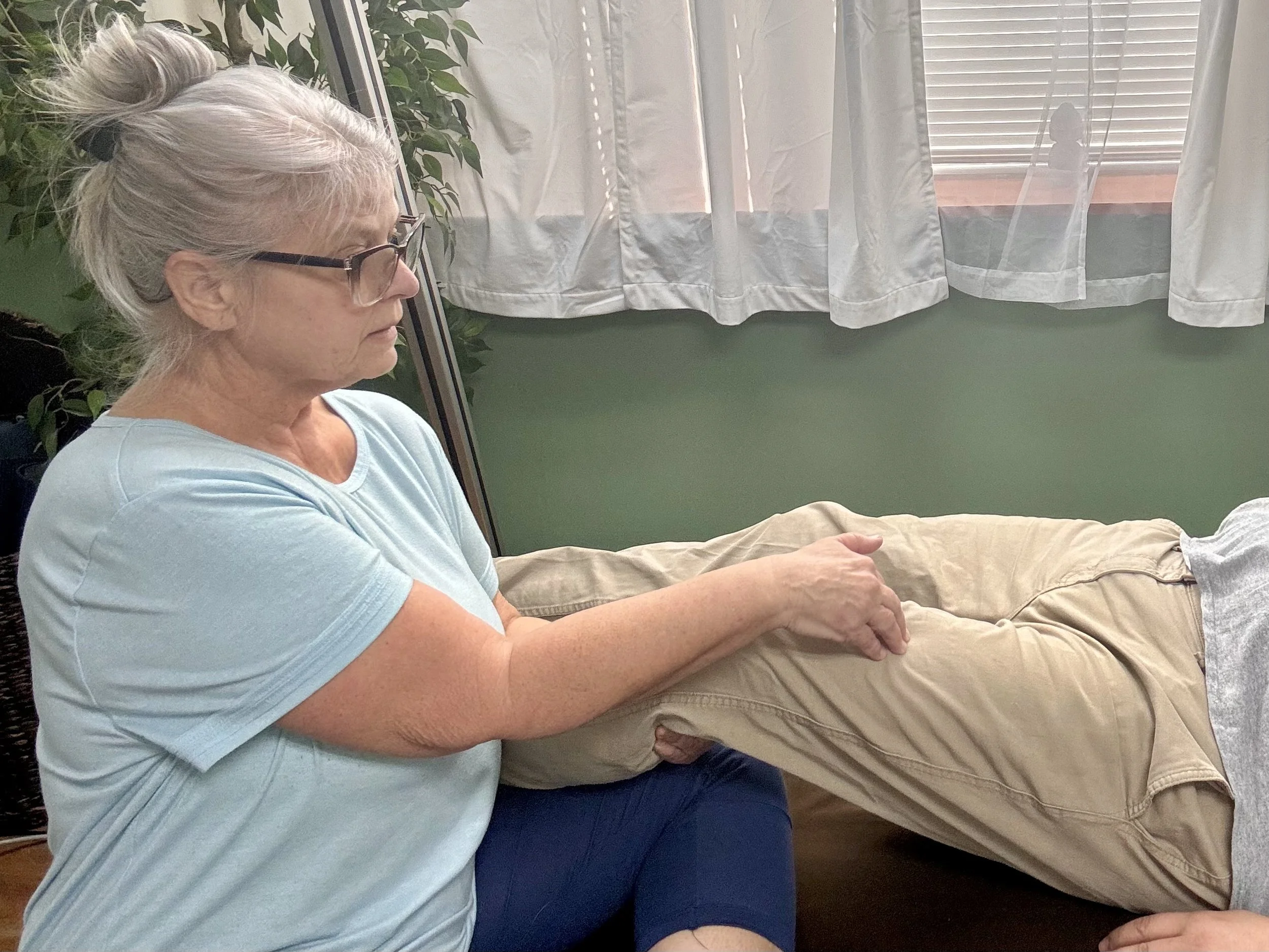 A woman with gray hair and glasses providing Zero Balancing to a person's leg, which is elevated and resting on her lap, in a room with green walls and white curtains.