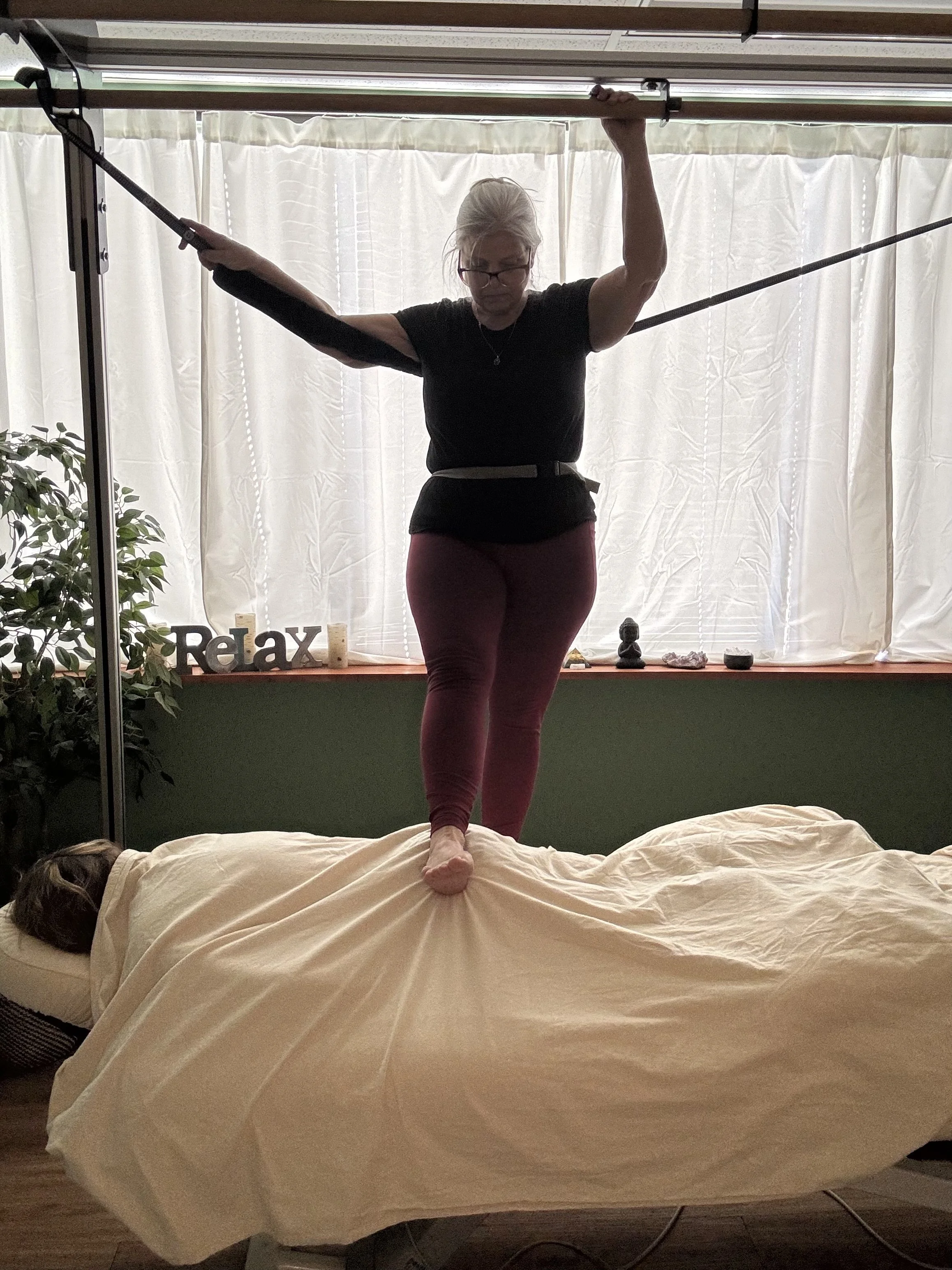 A woman practicing Ashiatsu massage on a massage table with a person lying down, in a room with white curtains, a plant, and decorative items on the windowsill spelling 'Relax'.