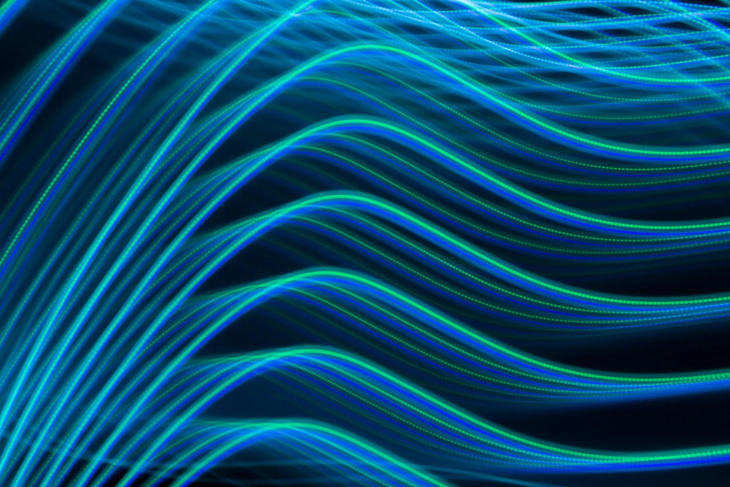 Ondes lumineuses ondulées bleues et vertes sur fond noir. Composition abstraite évoquant flux de données numériques, réseau fibre optique, connexion internet rapide et innovation technologique.