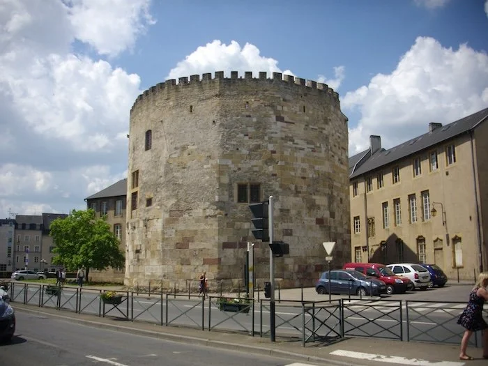 Tour fortifiée ronde en pierre, entourée de bâtiments résidentiels, avec des voitures garées et un ciel nuageux en arrière-plan.