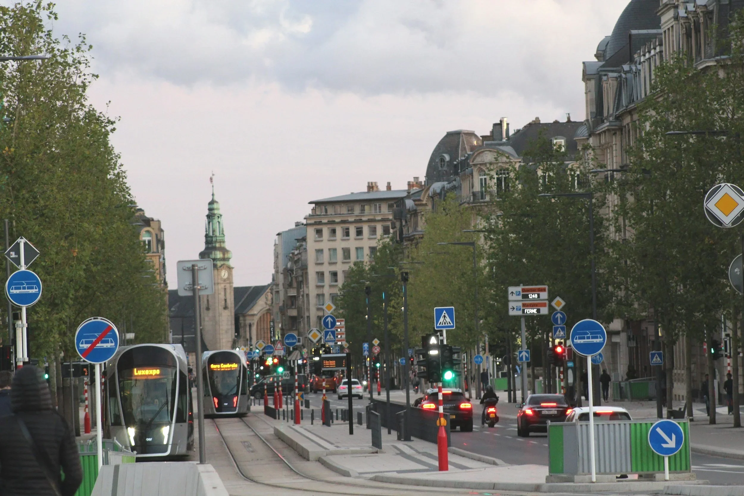 Tramways circulant sur voies ferrées de l'Avenue de la Liberté à Luxembourg-Ville. Voitures, signalisation urbaine et bâtiments historiques avec clocher en arrière-plan.