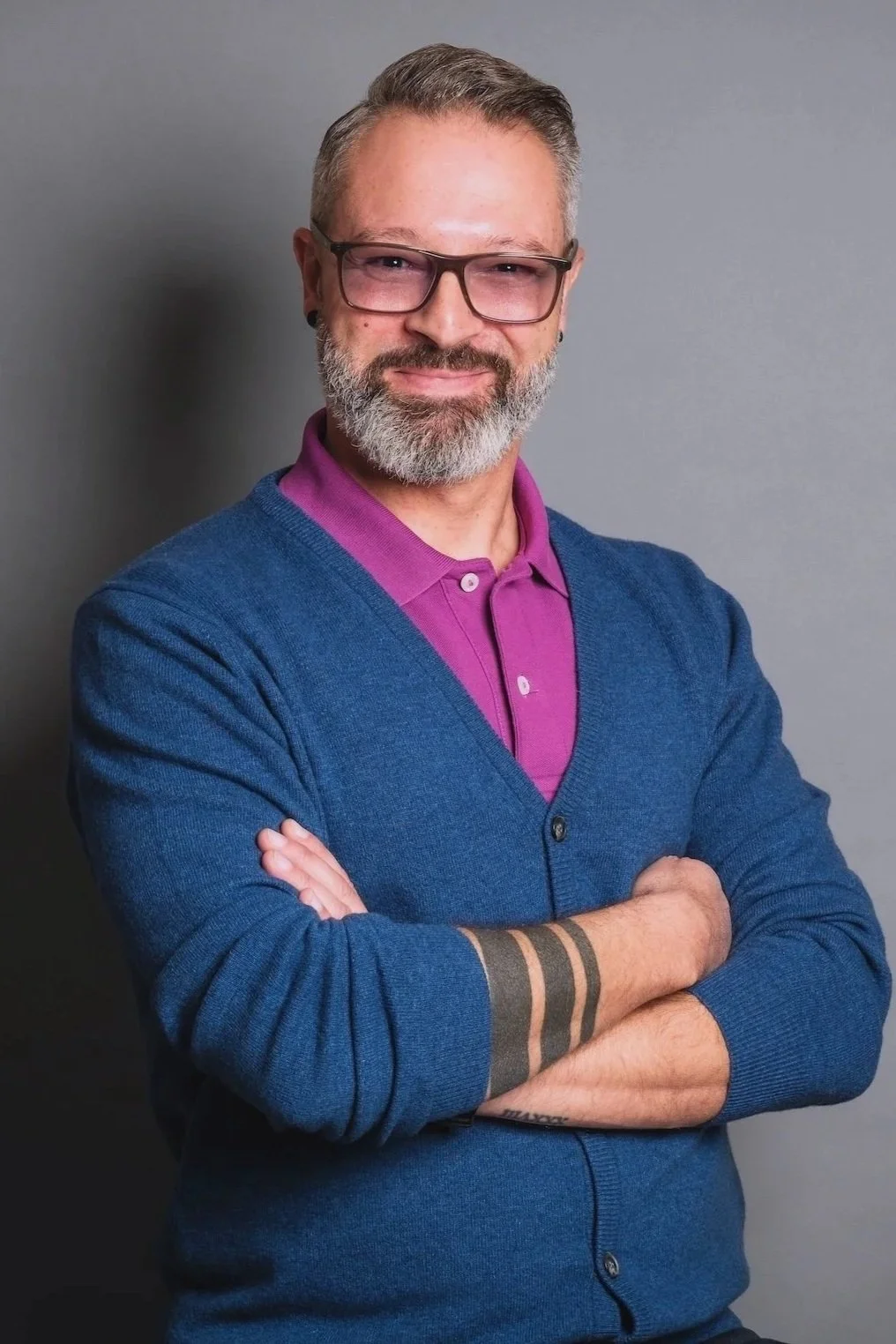 Portrait de Walid Karray souriant avec des lunettes, portant un pull bleu sur un polo violet, croisant les bras devant un mur gris.