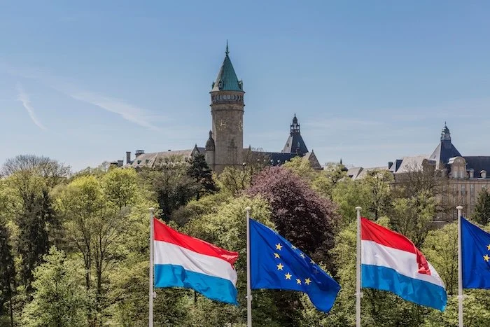 Quatre drapeaux, deux d'Autriche et deux de l'Union Européenne, flottent devant un château médiéval entouré d'arbres en fleurs peu après le printemps.