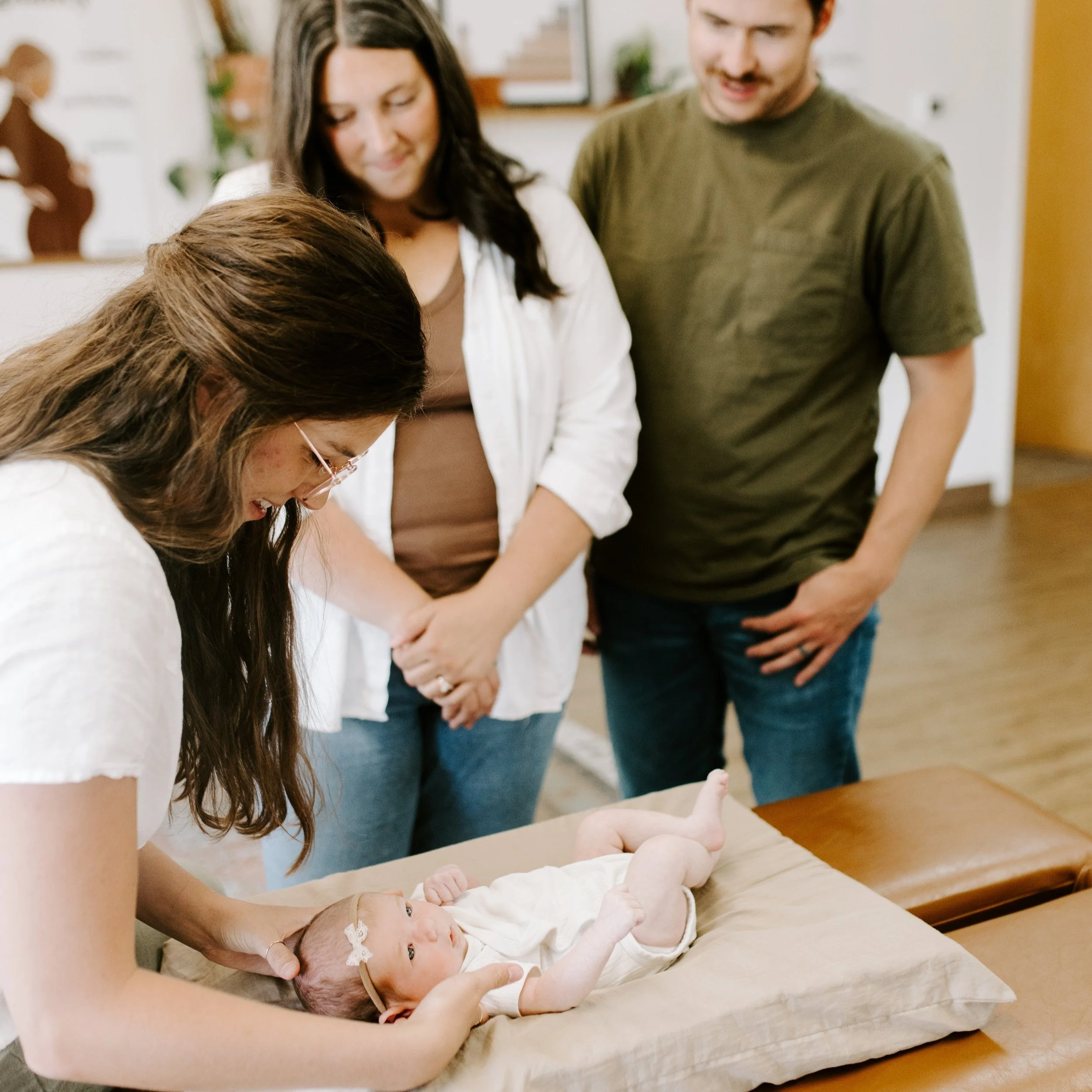 A woman with glasses holding a newborn baby on a changing table, while two adults look on affectionately in the background.