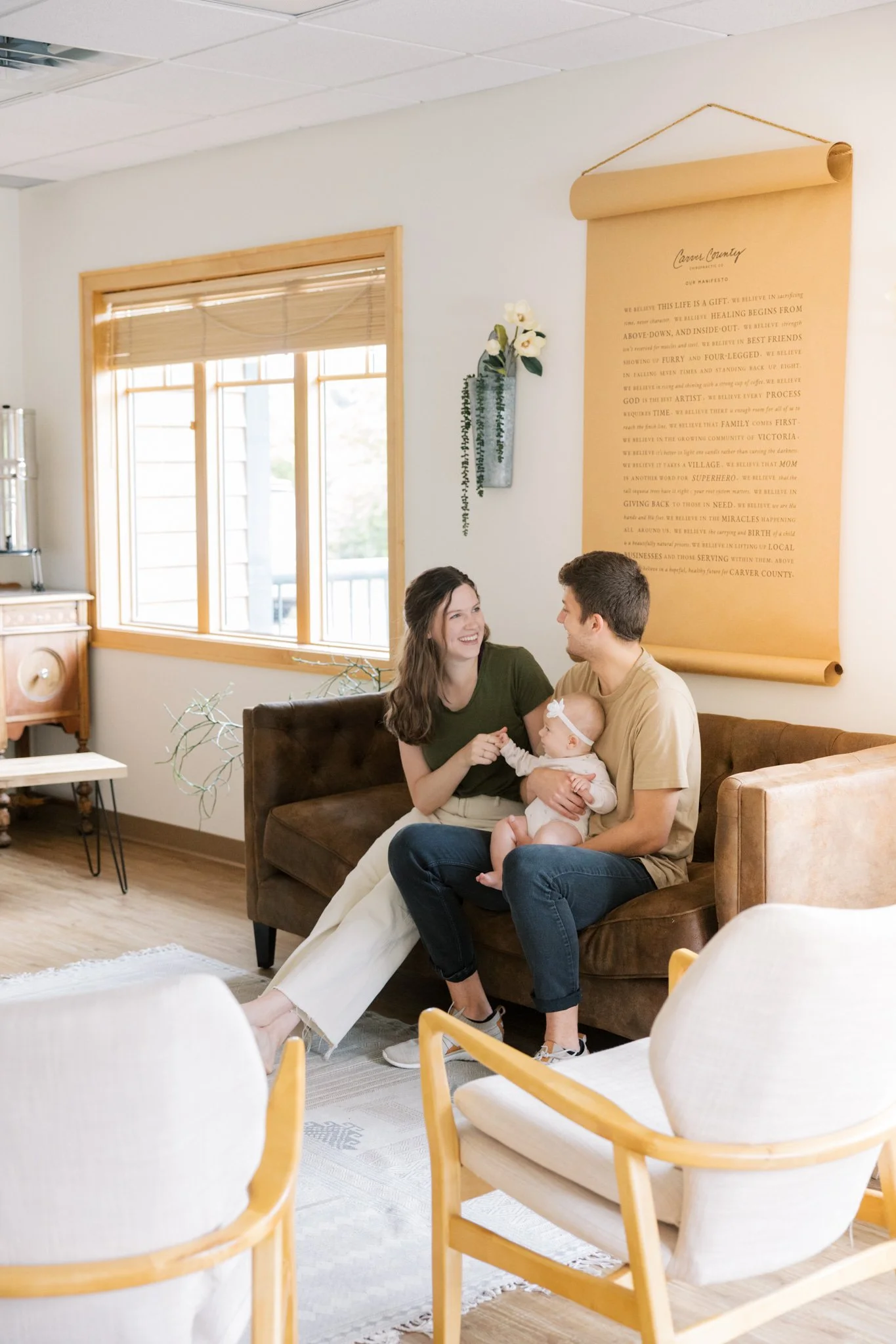 A couple sitting on a sofa holding a baby in a living room with large windows, wall decor, and modern furniture.