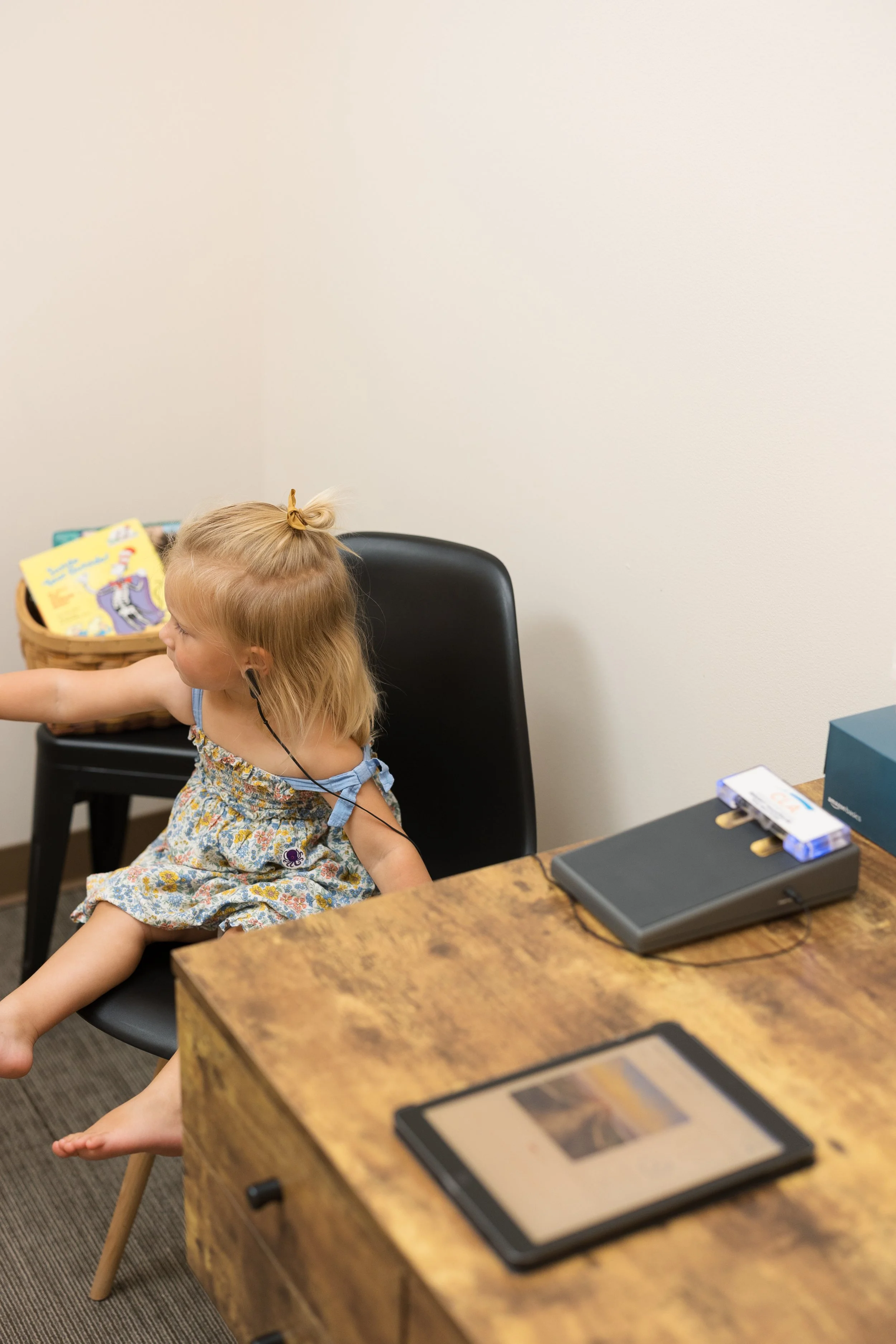 A young girl with blonde hair, a small ponytail with a yellow hairband, wearing a floral dress, sitting on a black chair in a room with a beige wall. There is a basket with a children's book and some items on a table nearby, and a device with cables and a small transparent plastic case on the table.