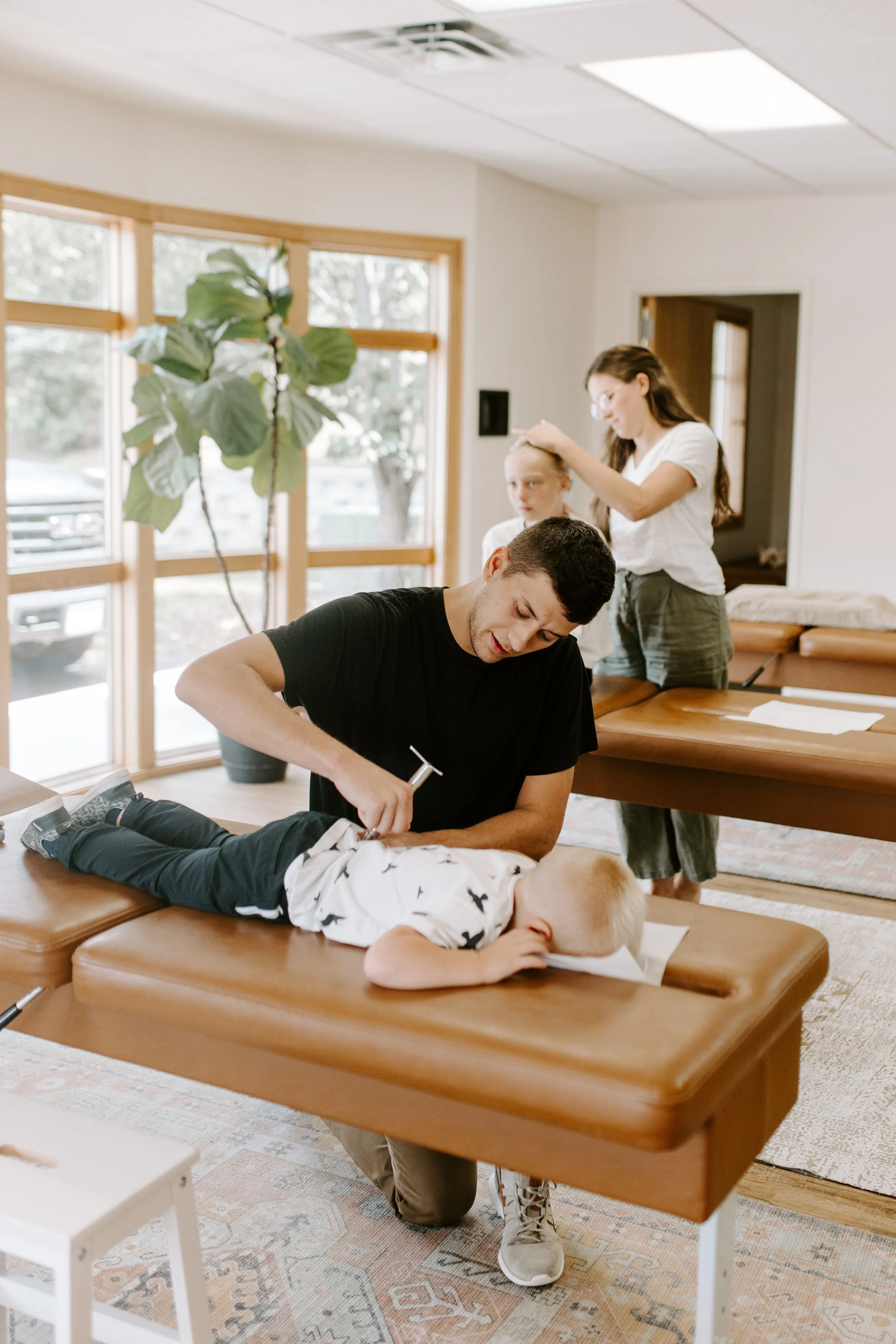 A man is lying face down on a chiropractic table with a child's head resting on table. A woman is adjusting a girl in the background, while another girl looks on. The scene is in a well-lit room with large windows and a potted plant.