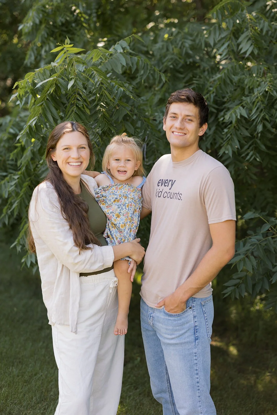 A smiling family of three standing outdoors in front of green trees. The mother holds a young girl, and the father stands beside them, all smiling.