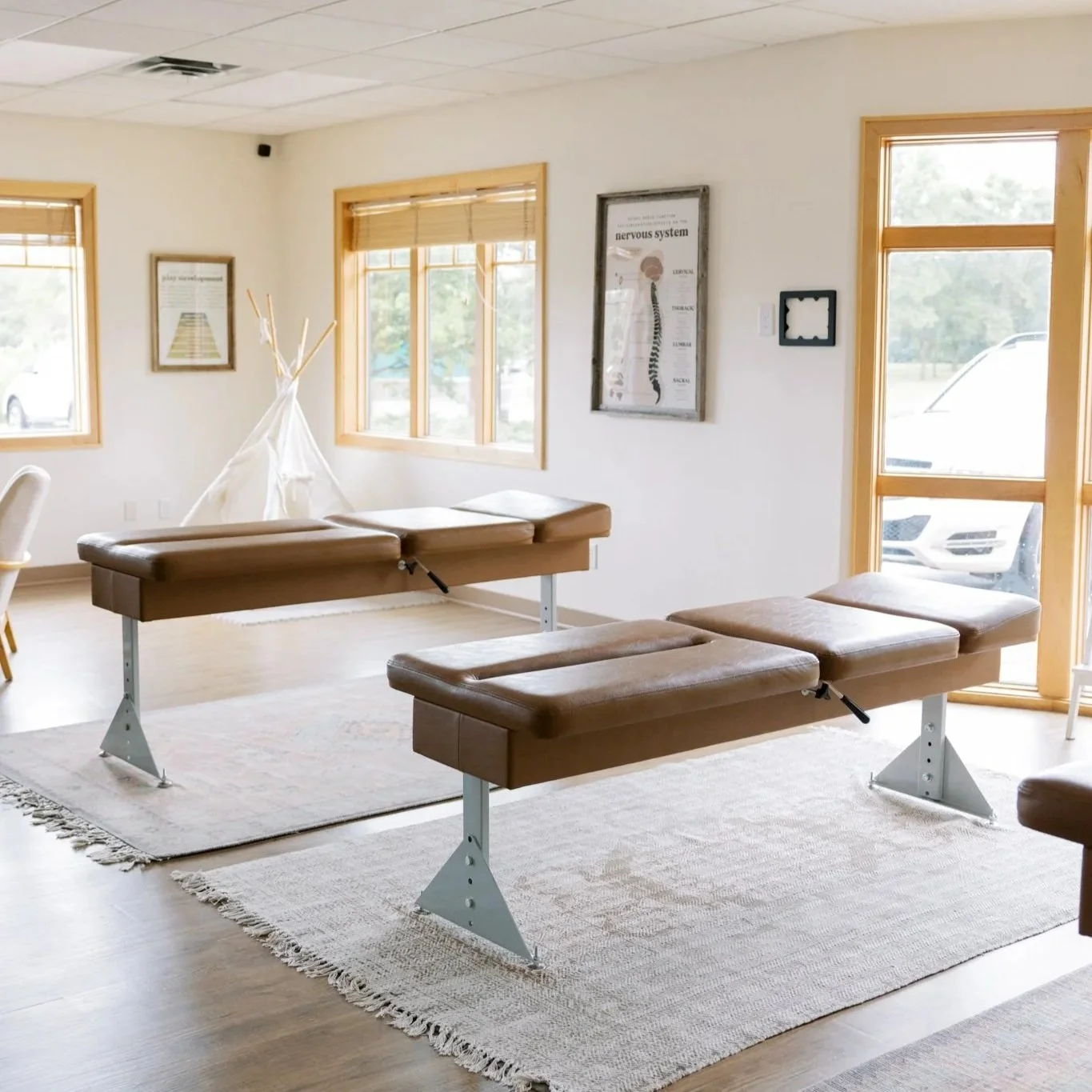 A chiropractic clinic room with two brown treatment tables on a beige rug, a white teepee in the corner, framed posters on the white walls, and windows with blinds allowing natural light.