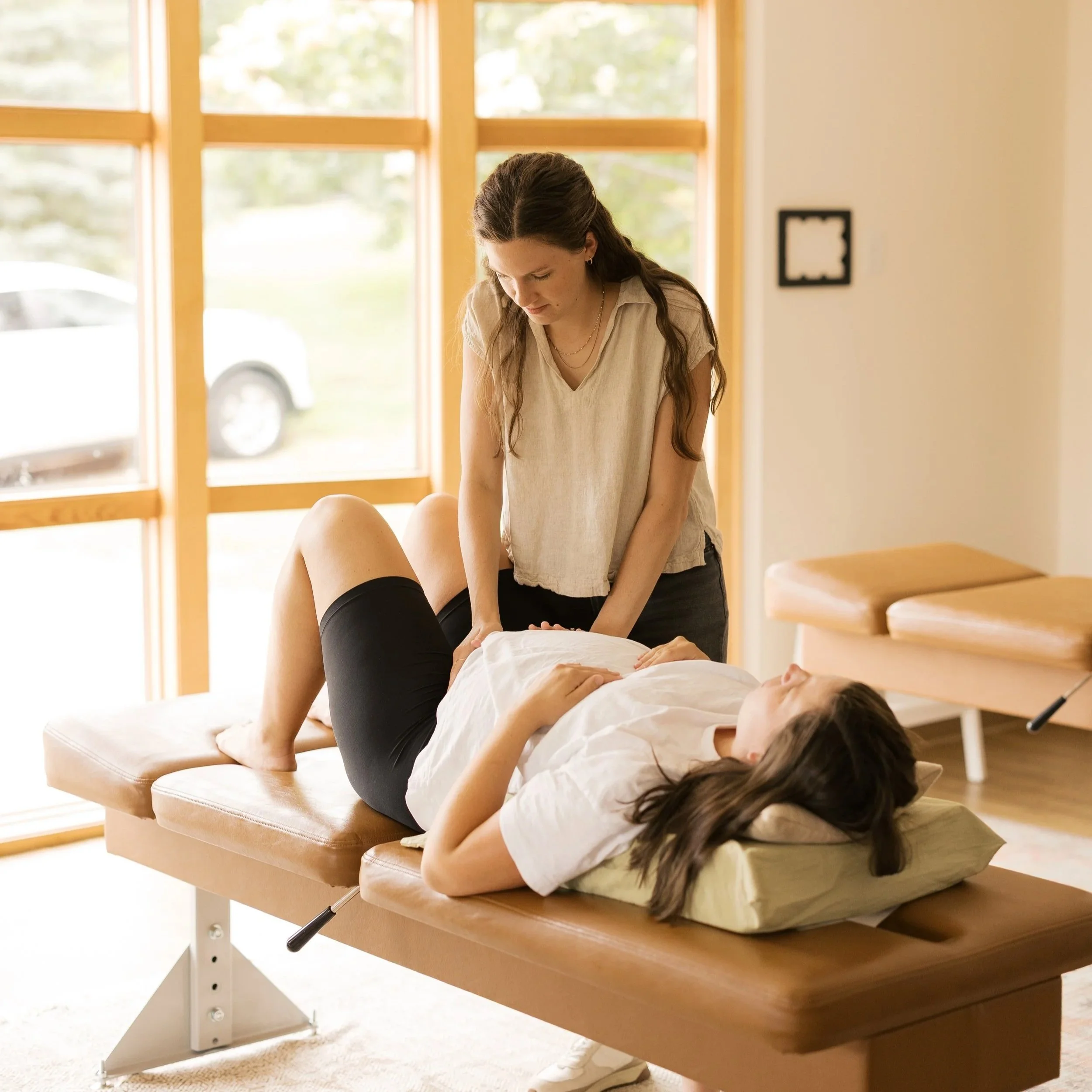 A woman in casual clothing performs chiropractic on a person lying face up on a chiropractic table in a bright room with large windows and wooden accents.