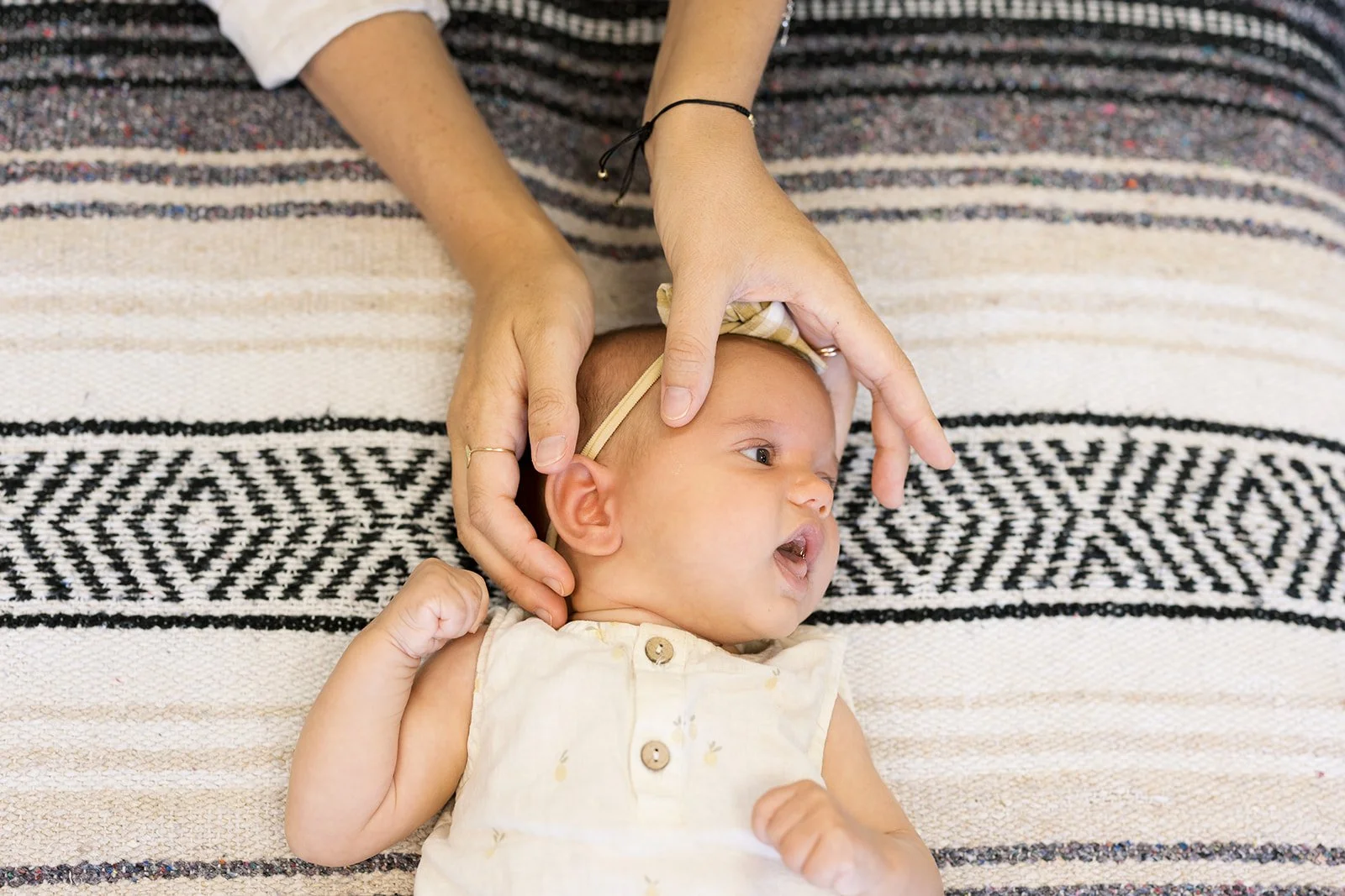 Close-up of a baby lying on a patterned blanket, being gently held and examined by an adult, with part of the adult's arm and hand visible.