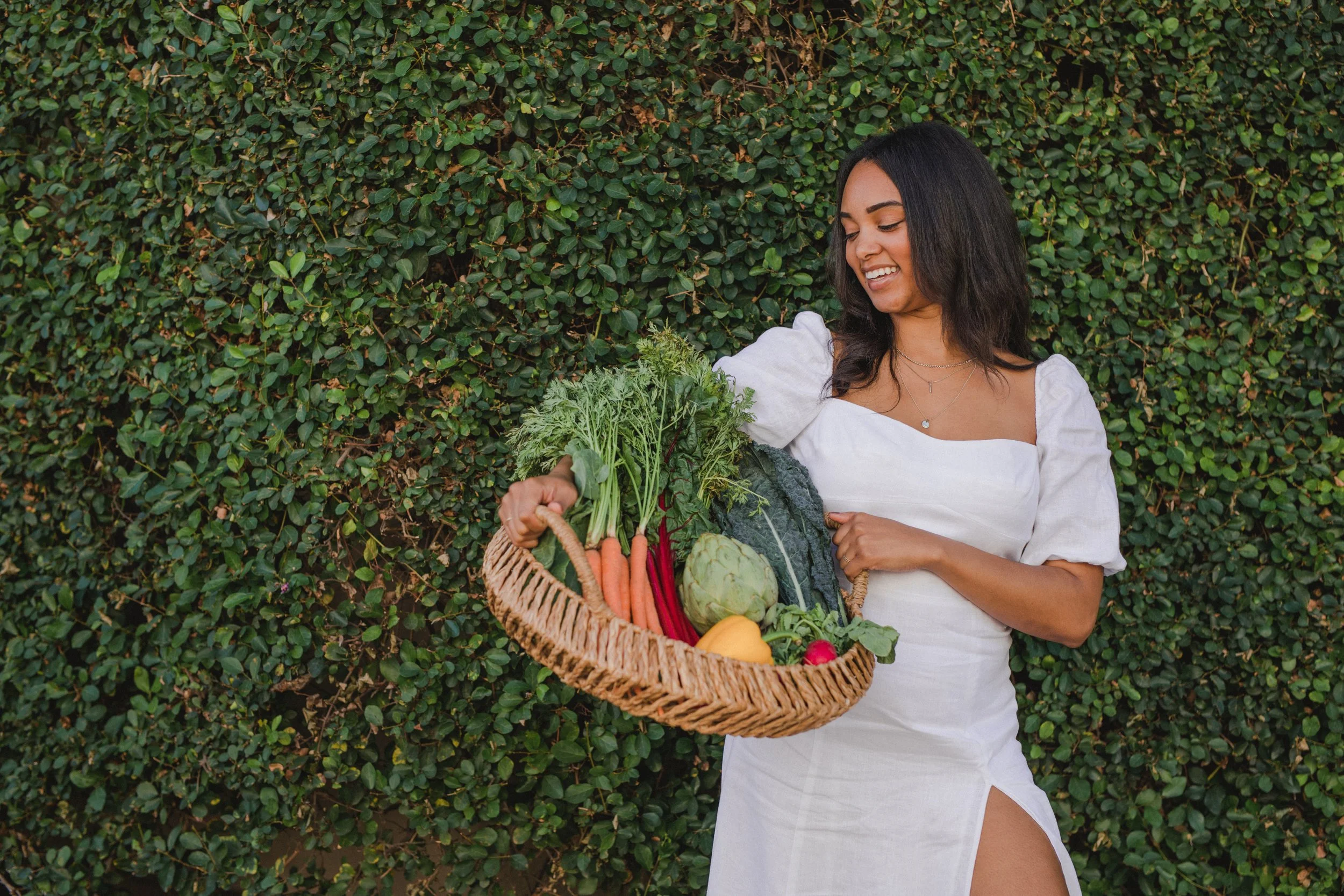 Image of Maya Amadi Howard, a 33 year old Private Chef, Writer, and Food Activist, wearing a white dress from Reformation, smiling while holding a wicker basket of fresh vegetables. She is standing in front of a wall of greenery.