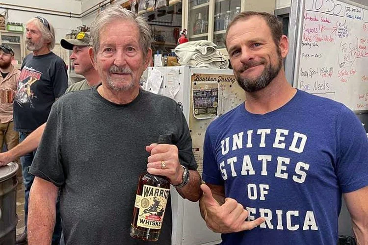 Nathan Newhall stands next to a man in a gray shirt holding a bottle of Warrior Whiskey while they tour the distilllery.